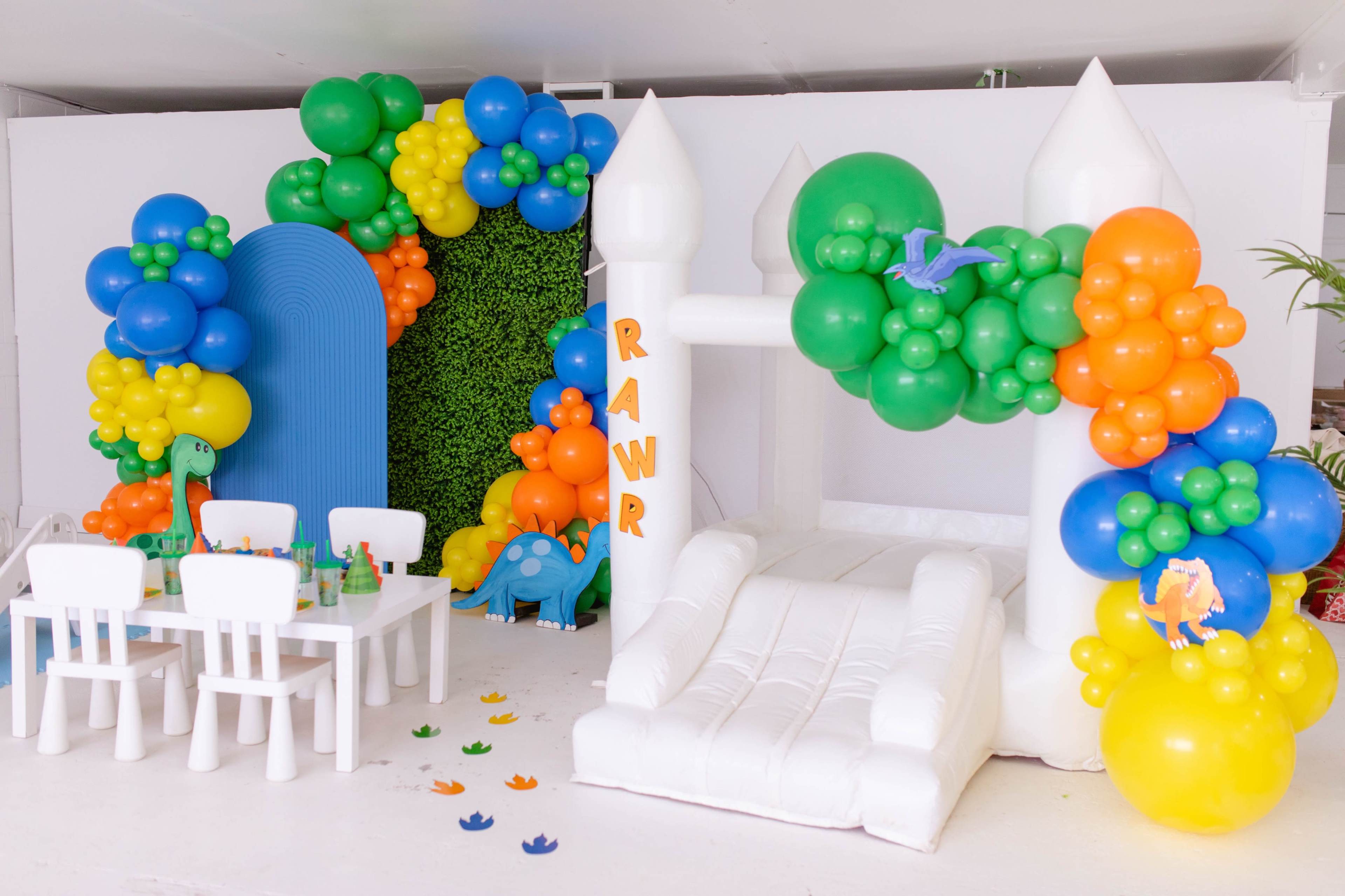 A vibrant party area featuring a white inflatable bounce house, decorated with colorful balloons in a dinosaur theme, alongside a small table set for children.