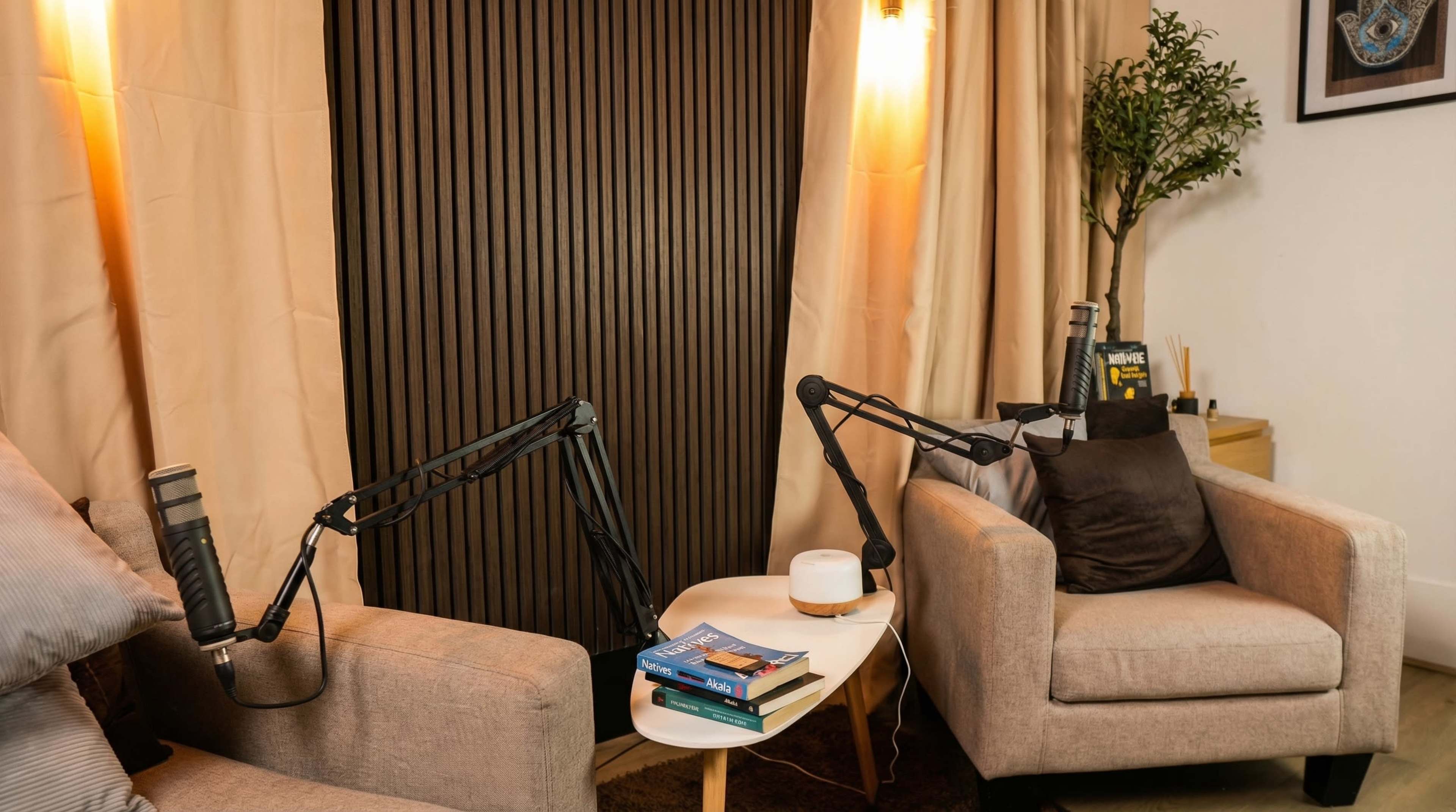 The image shows a small recording studio setup with two armchairs, a coffee table stacked with books, and two microphones mounted on flexible arms.