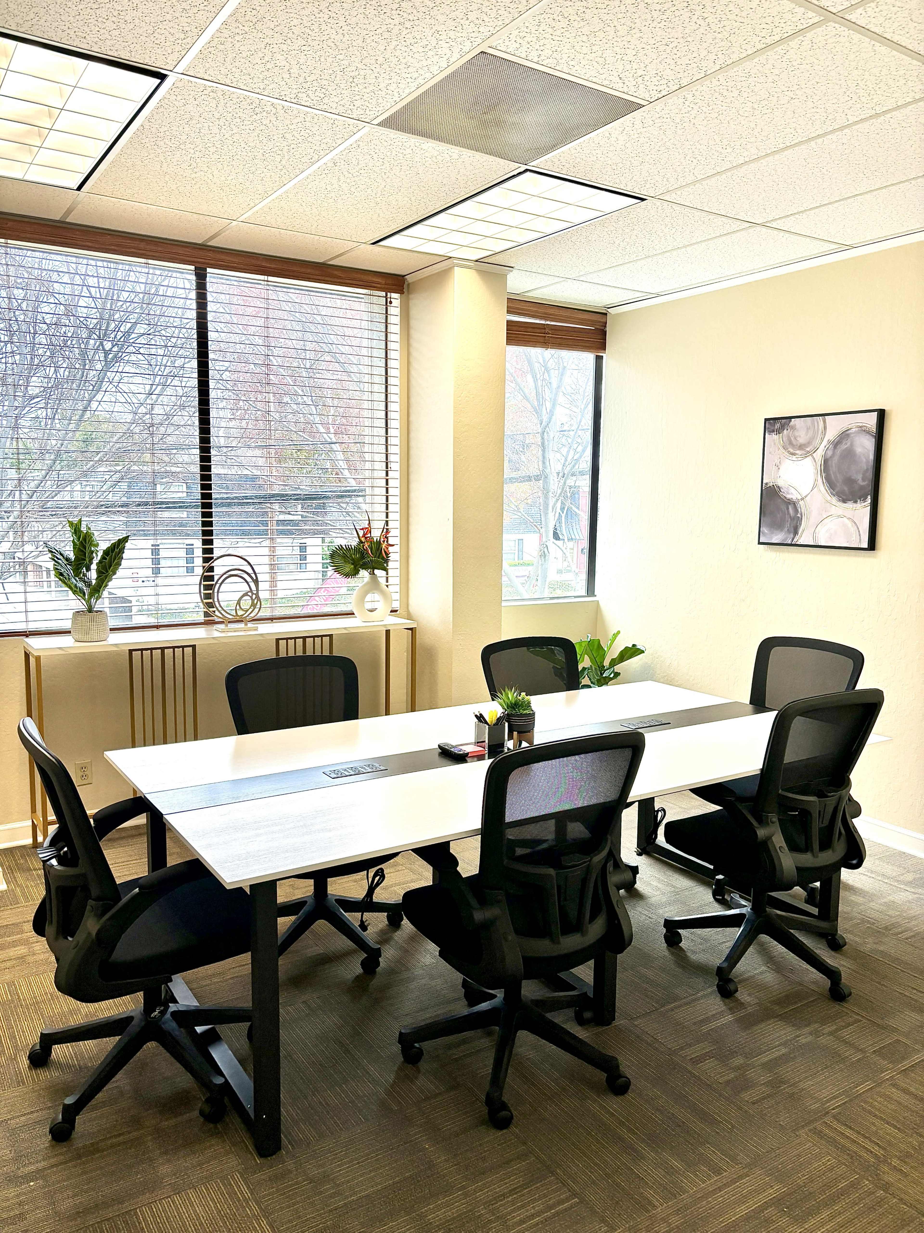 A bright office meeting room features a large white table surrounded by four black chairs, with windows offering a view of outside greenery.