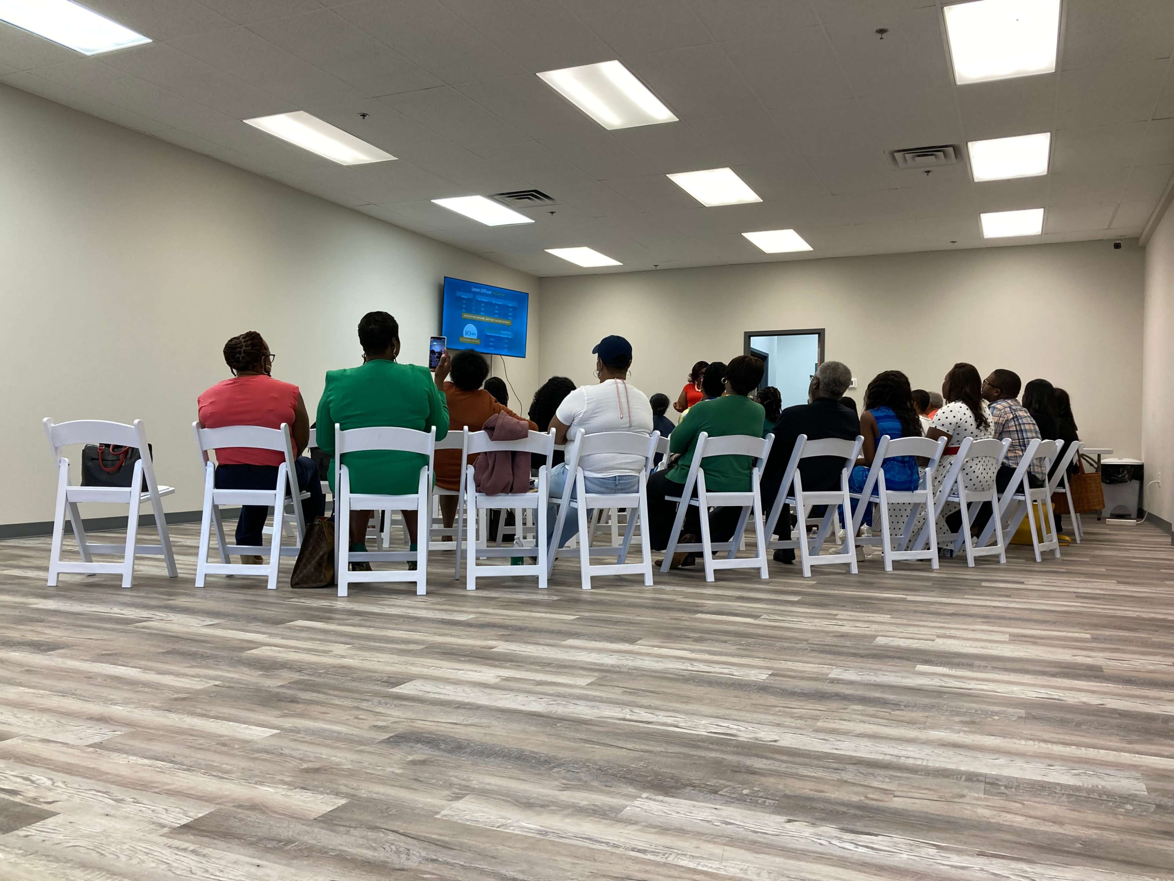 A group of people sit in rows of white chairs facing a screen in a plain room.