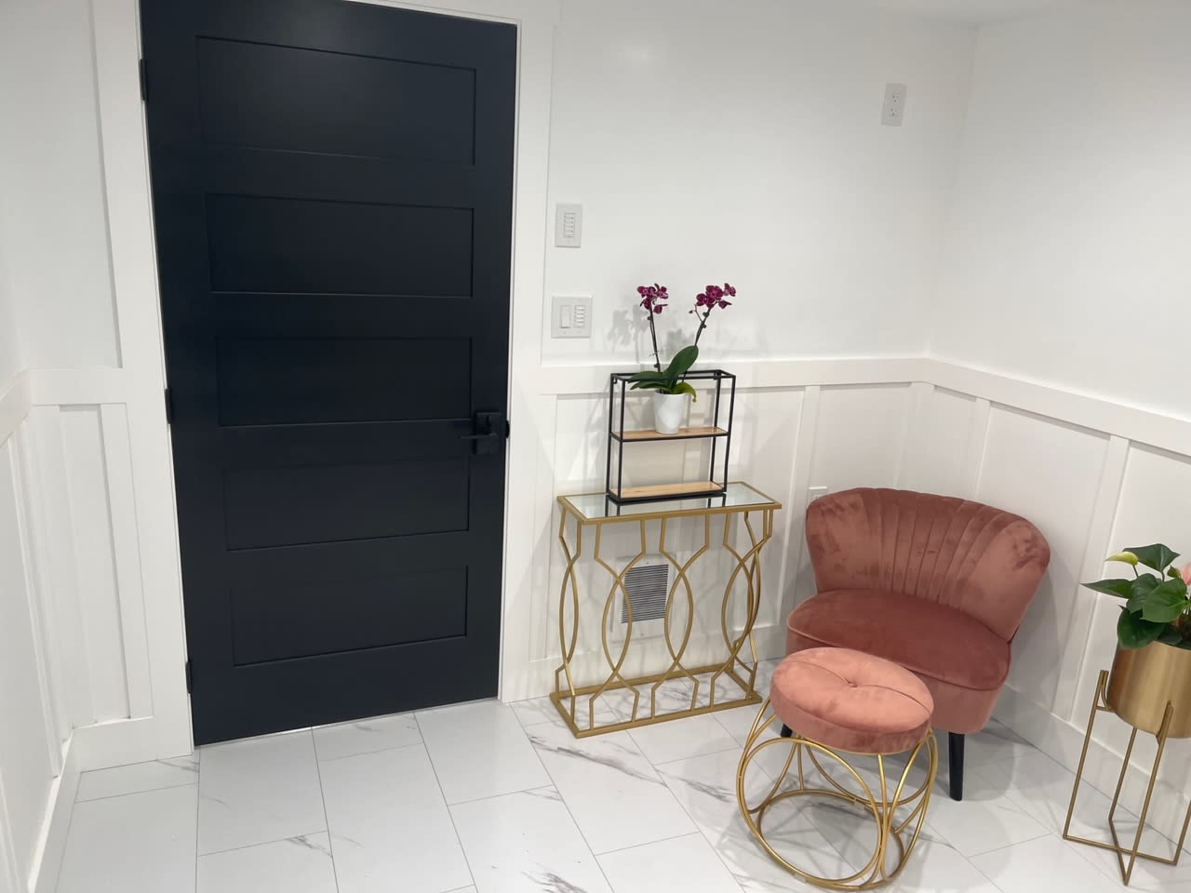 A modern entryway featuring a dark door, a pink upholstered chair, a gold framed console table, and potted plants against a white wall.