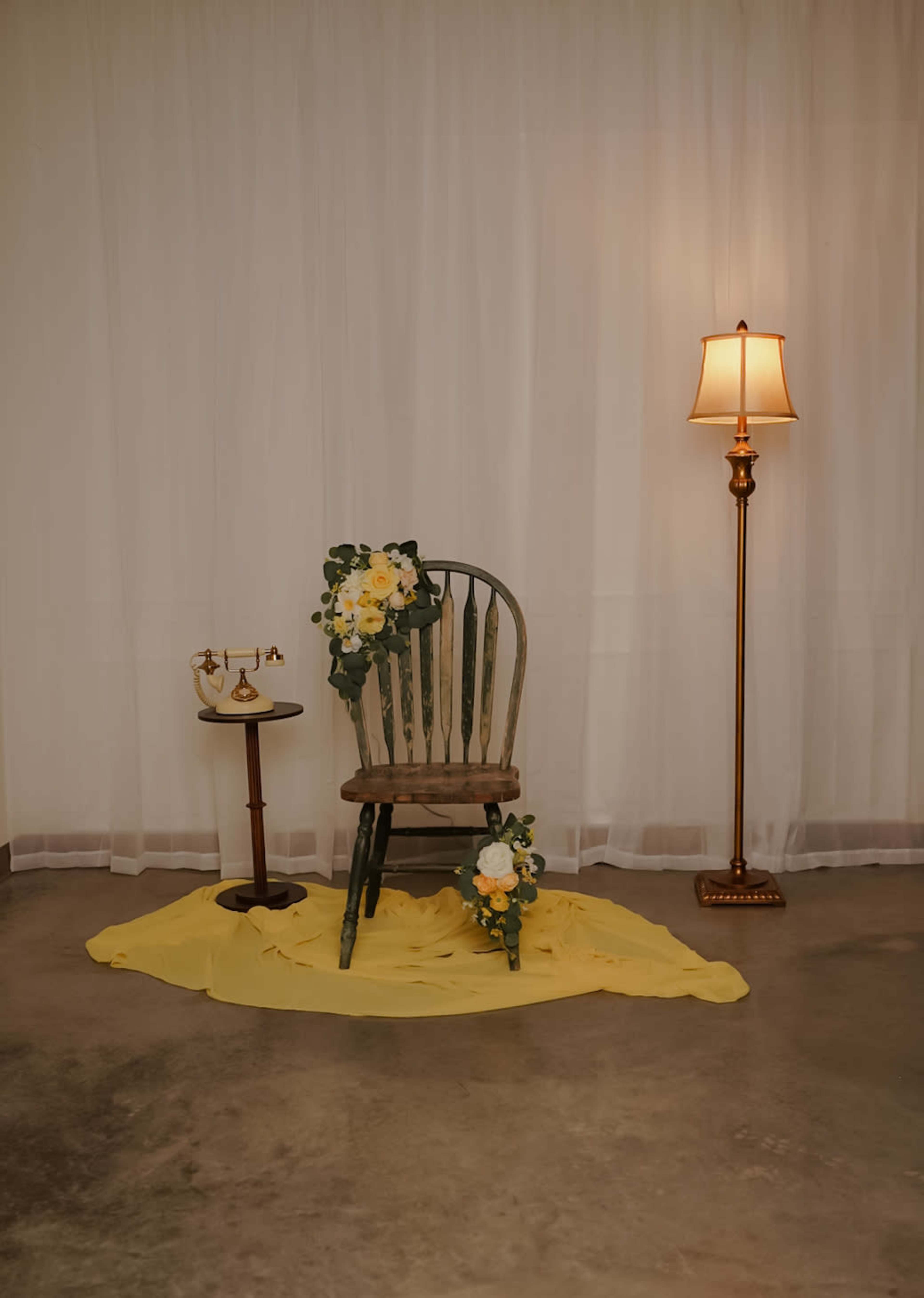A wooden chair with flowers sits on a yellow cloth beside a small table and a lamp in a softly lit room.
