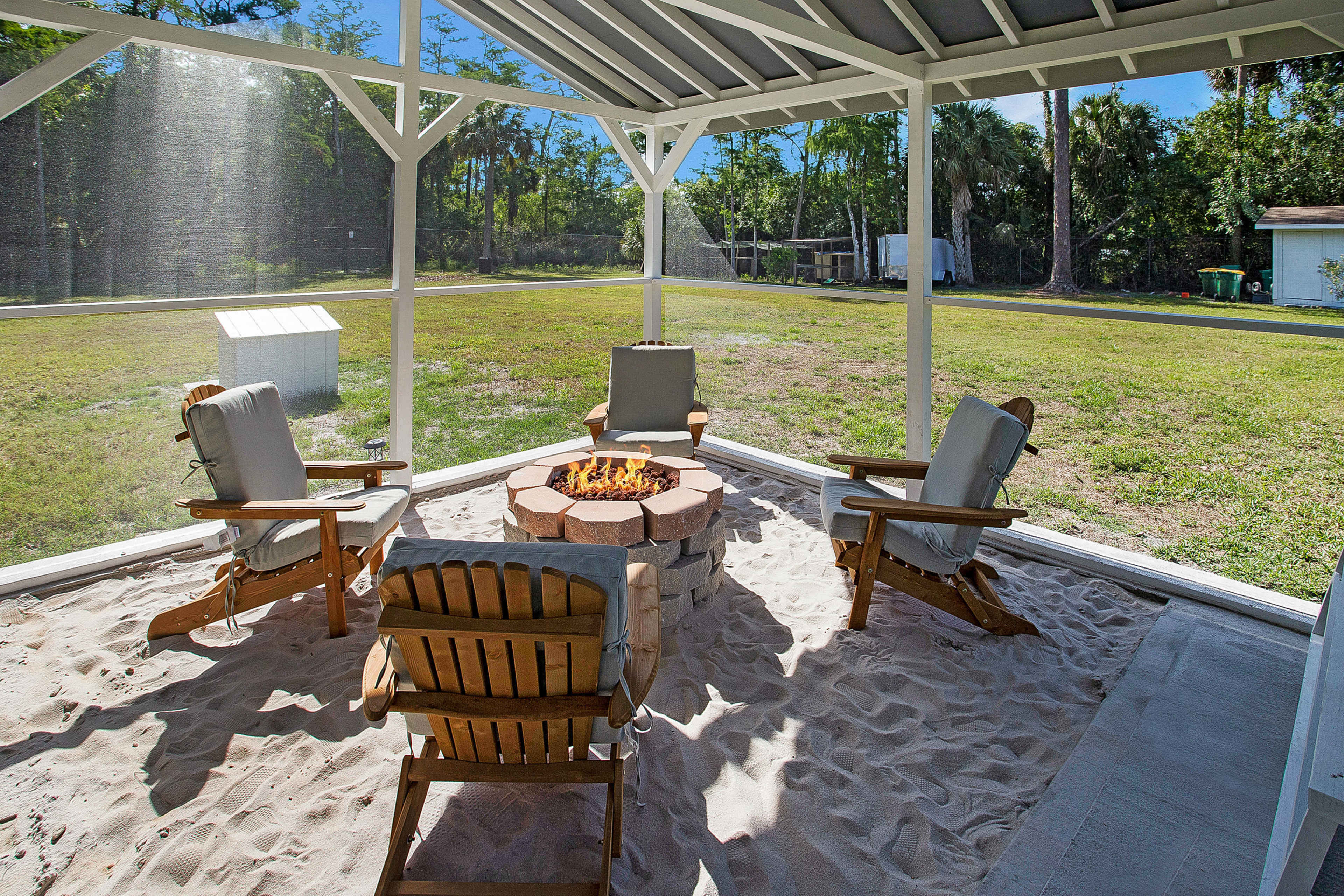 A screened-in outdoor area with a fire pit surrounded by three wooden chairs on sandy ground.