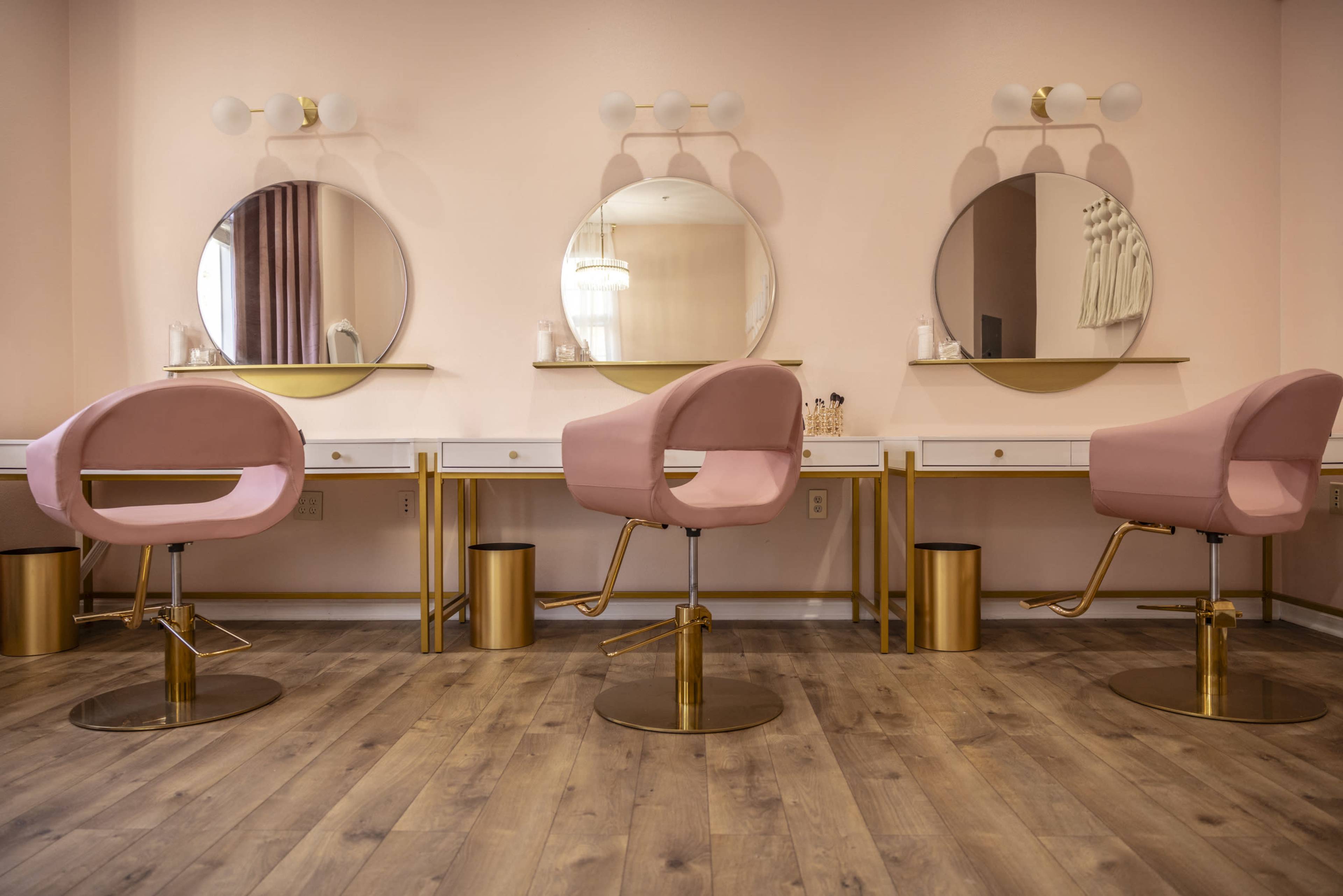 A salon interior with three pink upholstered chairs positioned in front of round mirrors on a light pink wall, complemented by gold accents and wooden flooring.