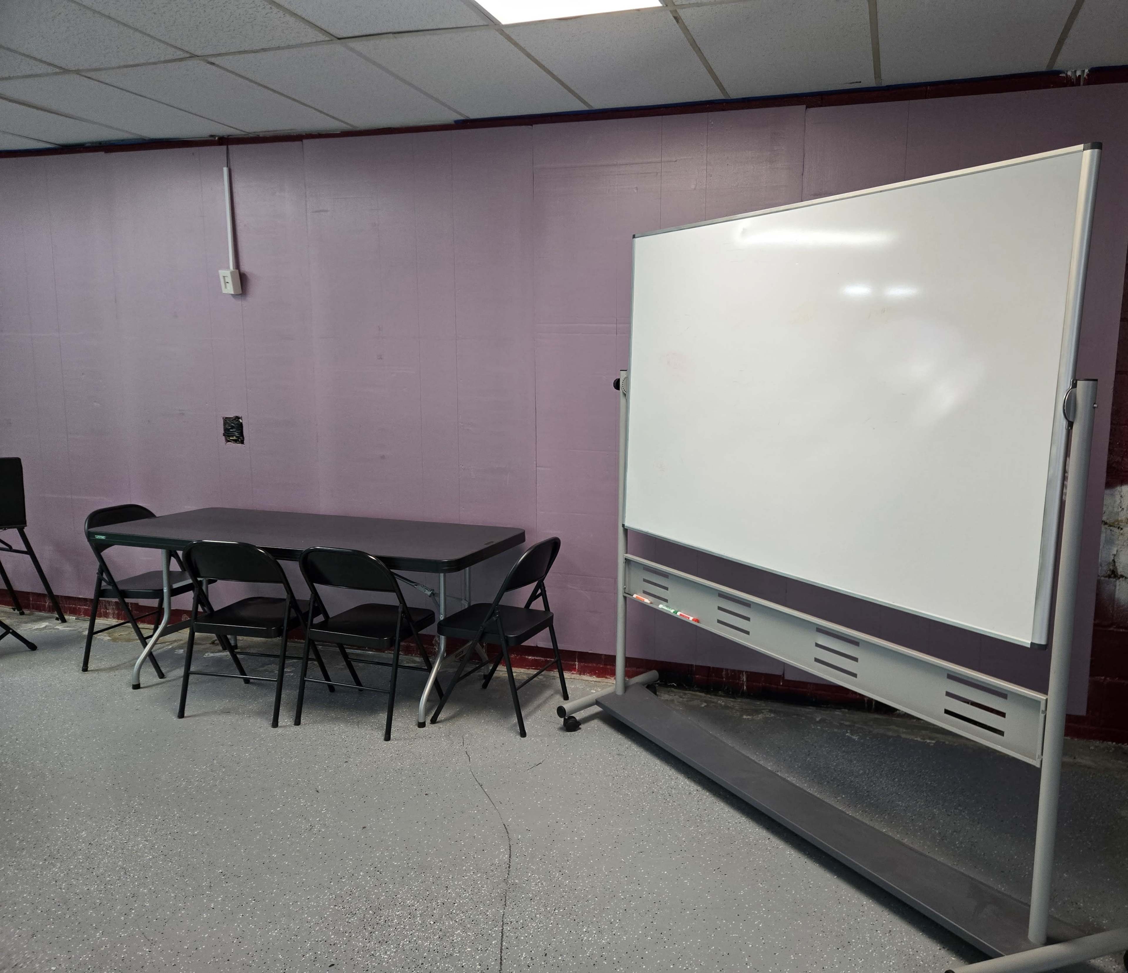 The image shows a room with a purple wall featuring a table with black chairs and a whiteboard on wheels.