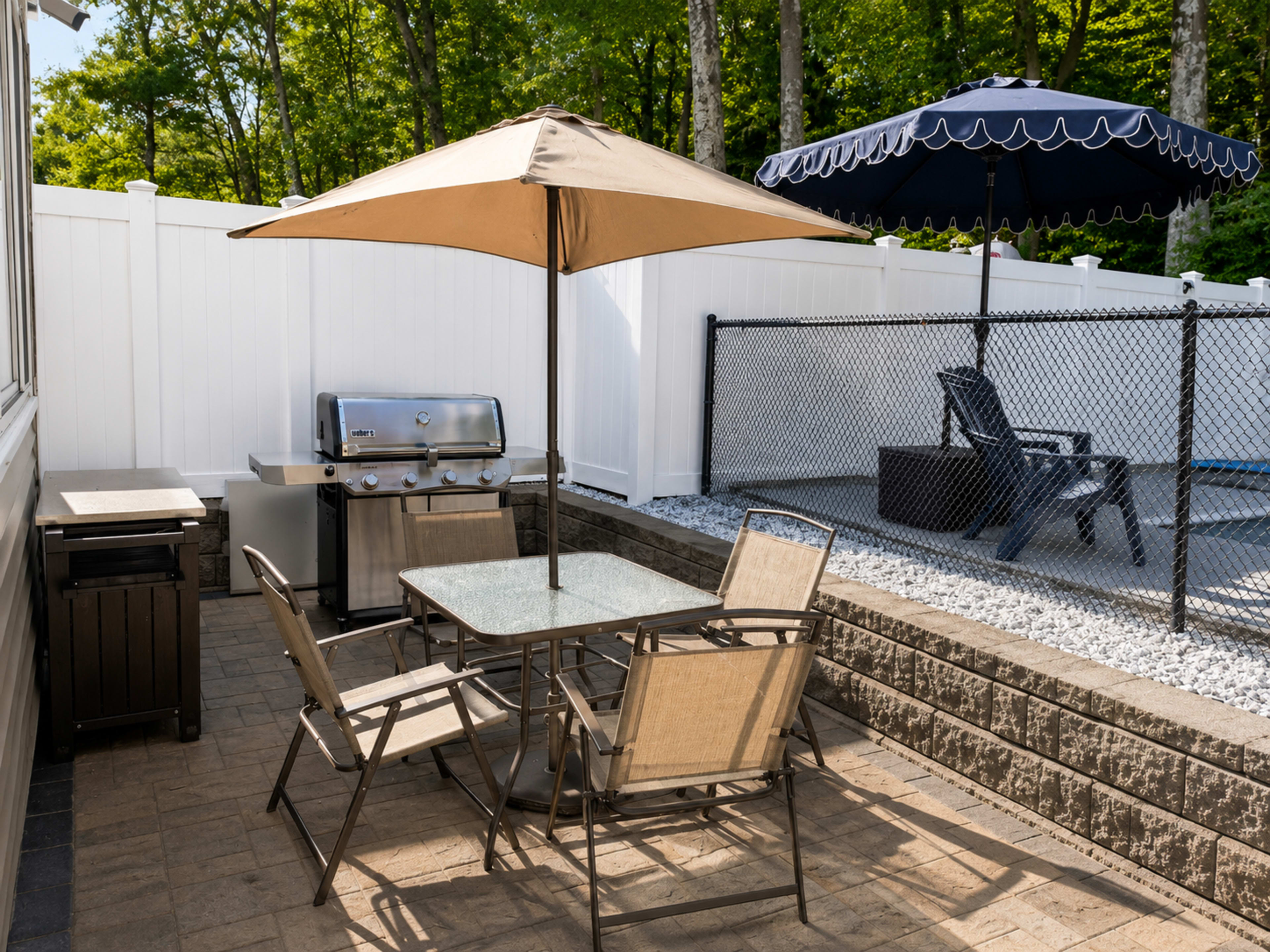 A patio area features a table with four chairs, a grill, and an umbrella, adjacent to a fenced-in space with lounge chairs.