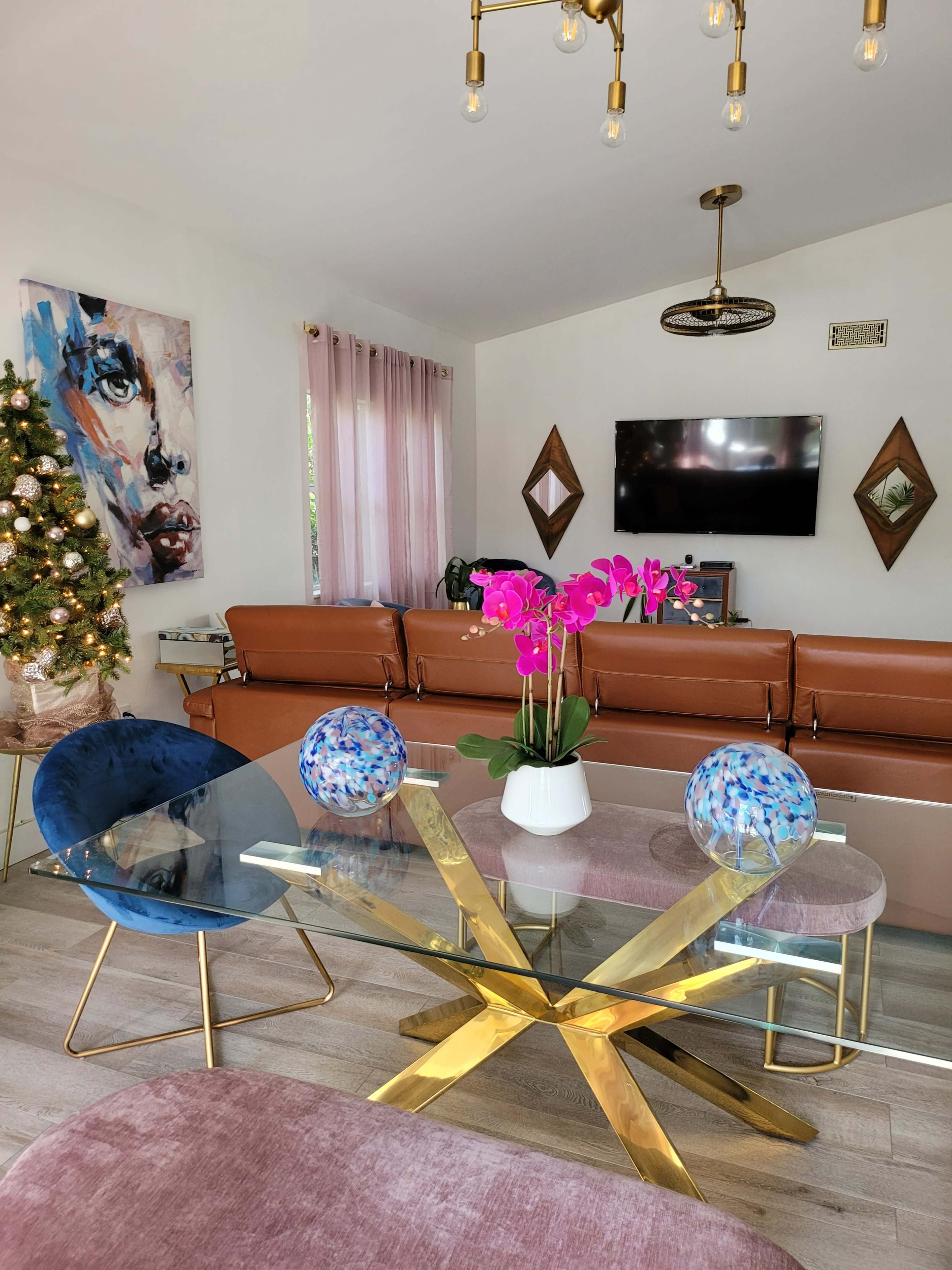 A modern living room features a glass dining table with gold accents, blue and pink decor, a flat-screen TV, and a decorated Christmas tree.