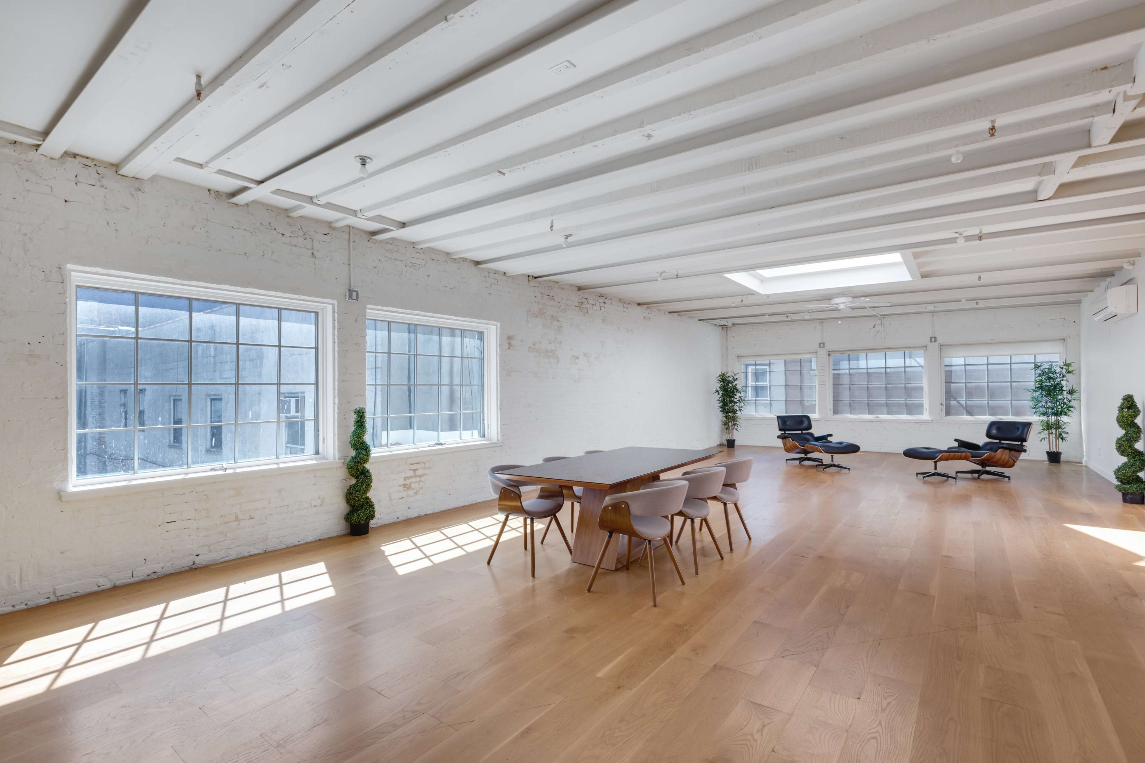 A spacious, brightly lit room features a wooden dining table surrounded by chairs, with large windows and minimalist decor, including potted plants and an office chair in one corner.