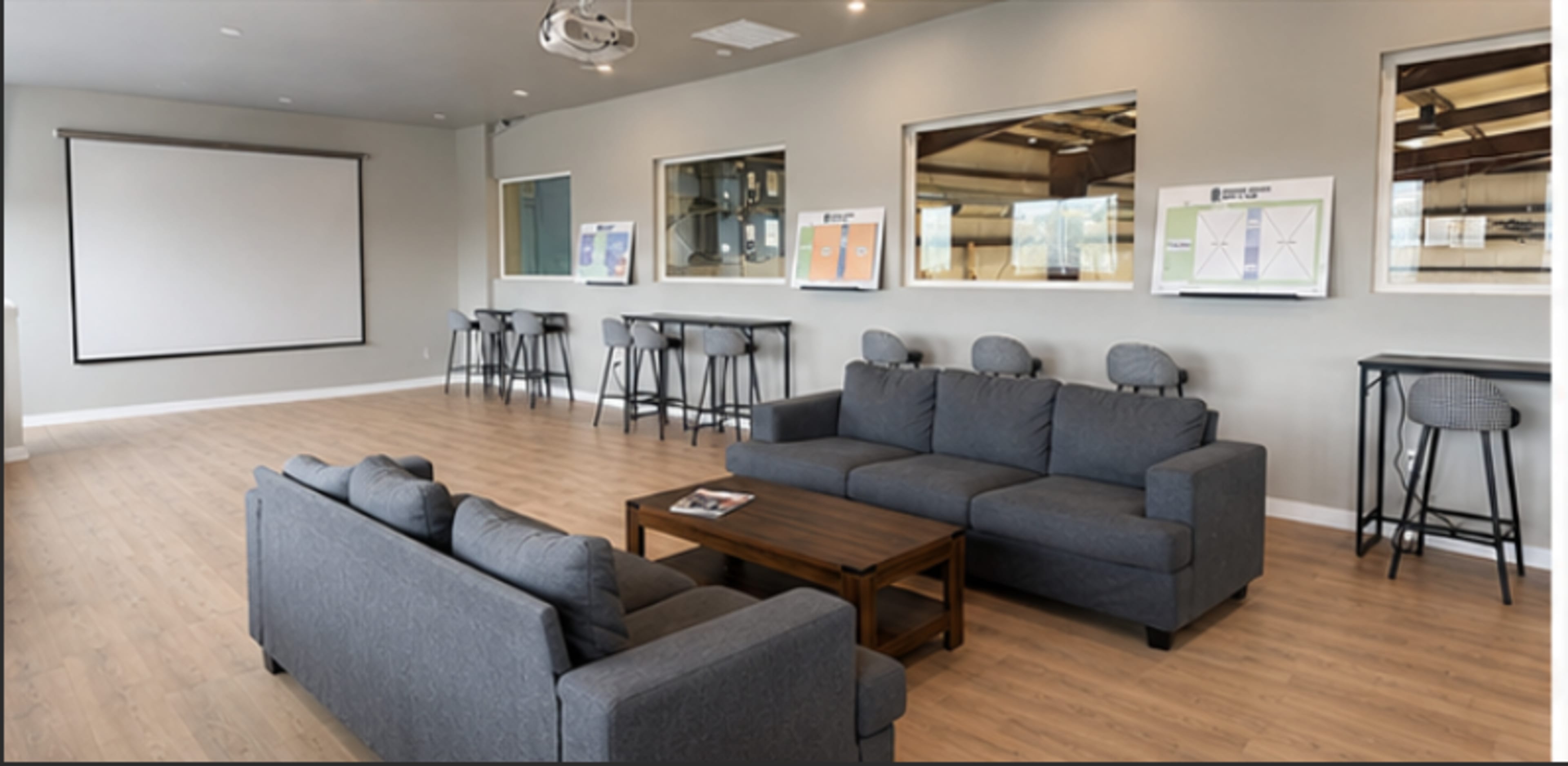 The image shows a modern meeting room with two gray sofas, a wooden coffee table, and a large blank projection screen mounted on the wall, alongside several framed displays.