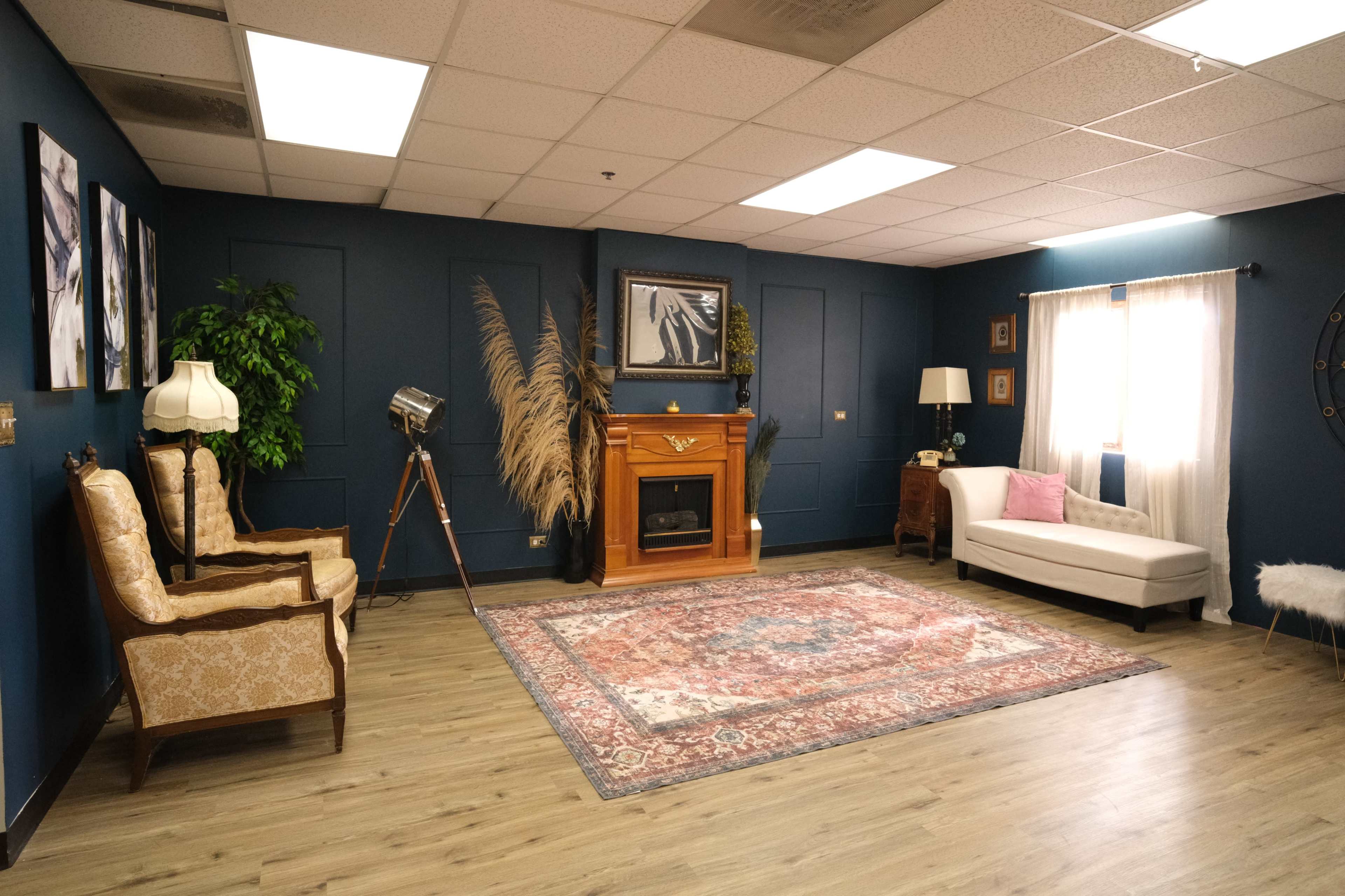 The room features a stylish seating area with two armchairs, a white sofa, a decorative fireplace, and a large rug, all set against dark blue walls.