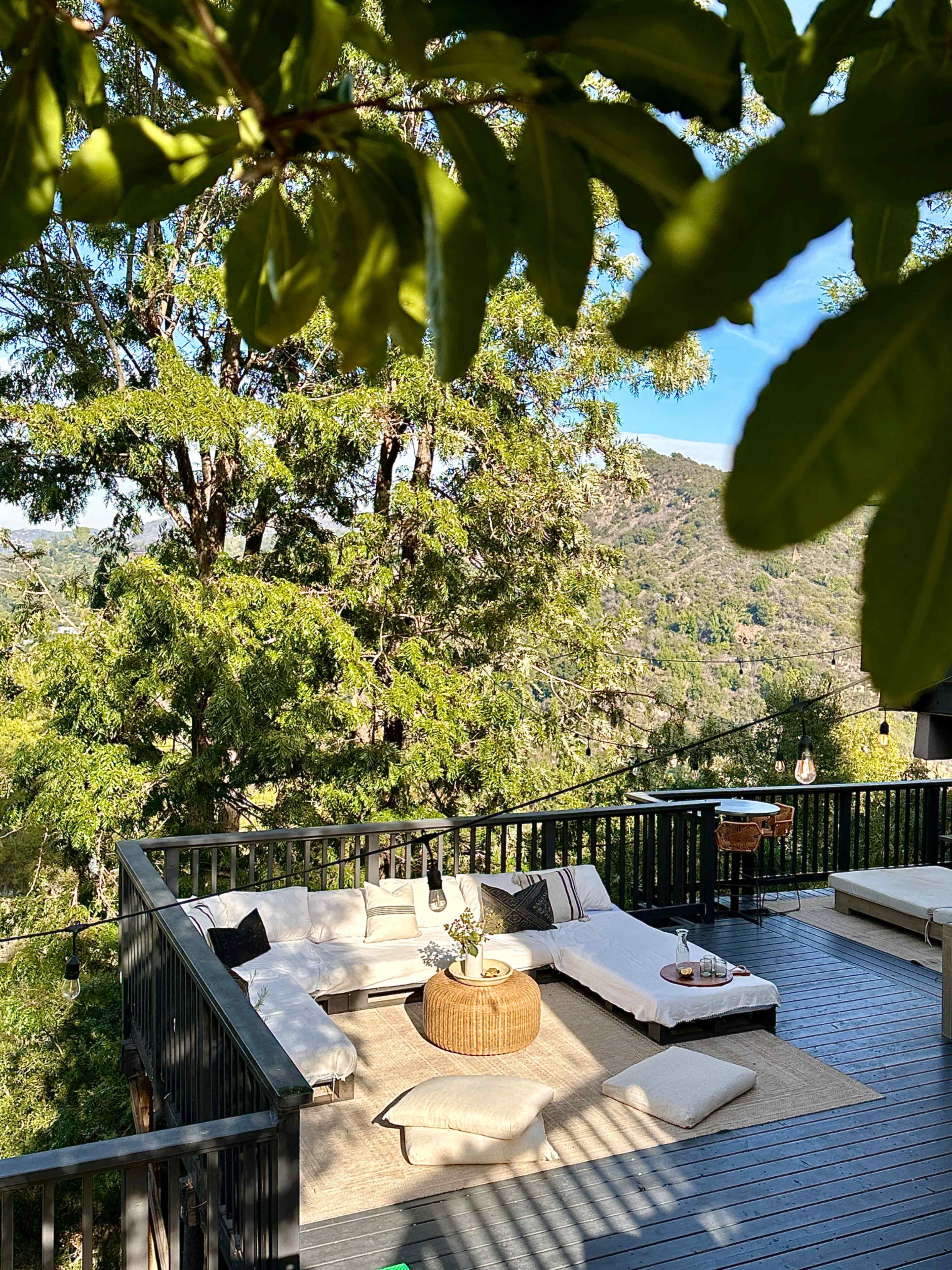 A spacious outdoor seating area features cushioned lounges and a round coffee table, surrounded by lush trees and mountainous scenery.