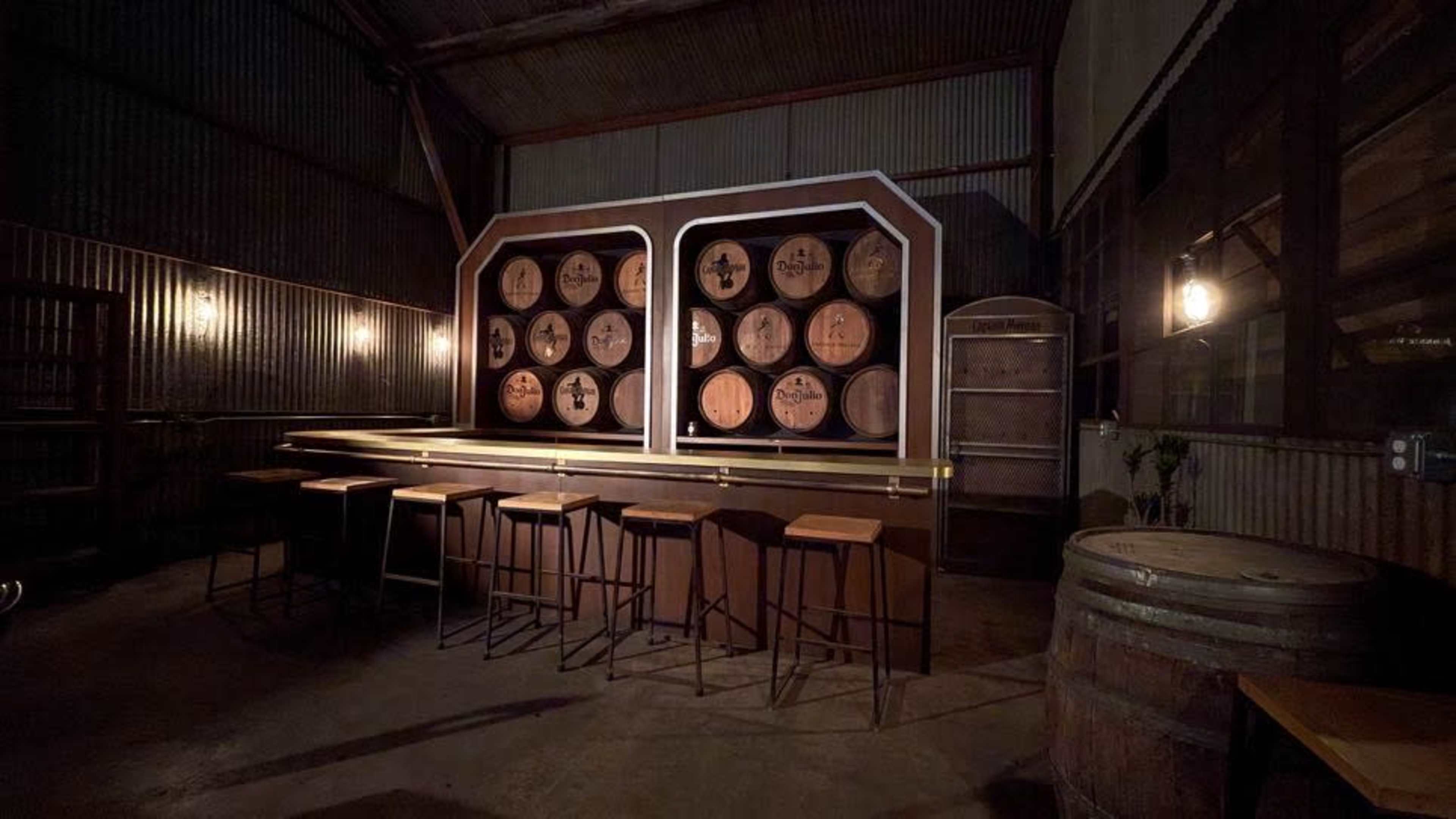 The image shows a dimly lit bar area with wooden barrels and a backdrop of circular wooden casks displayed on a wall.