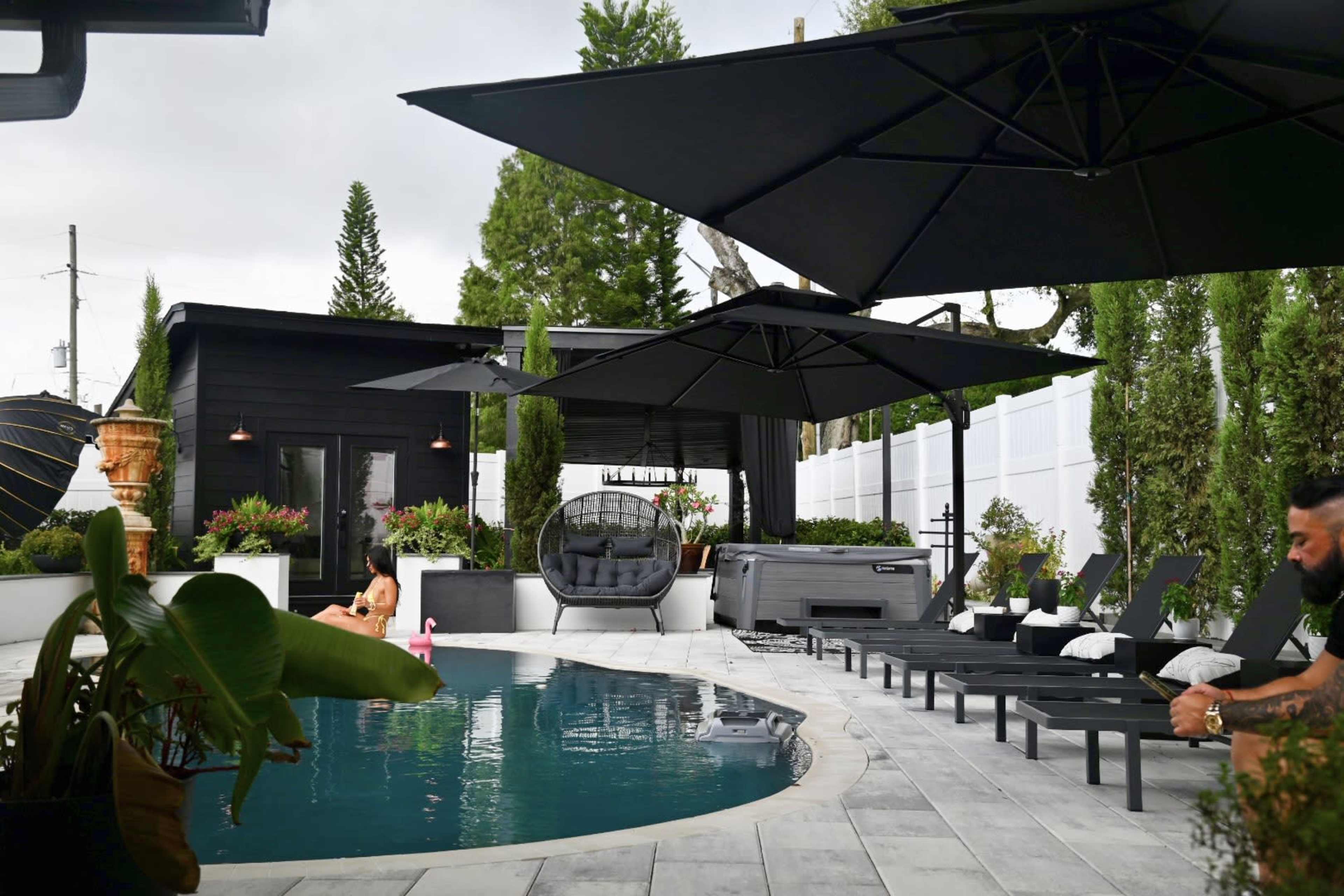 The image shows a modern outdoor space featuring a swimming pool, lounge chairs, large umbrellas, and a small building, surrounded by greenery.