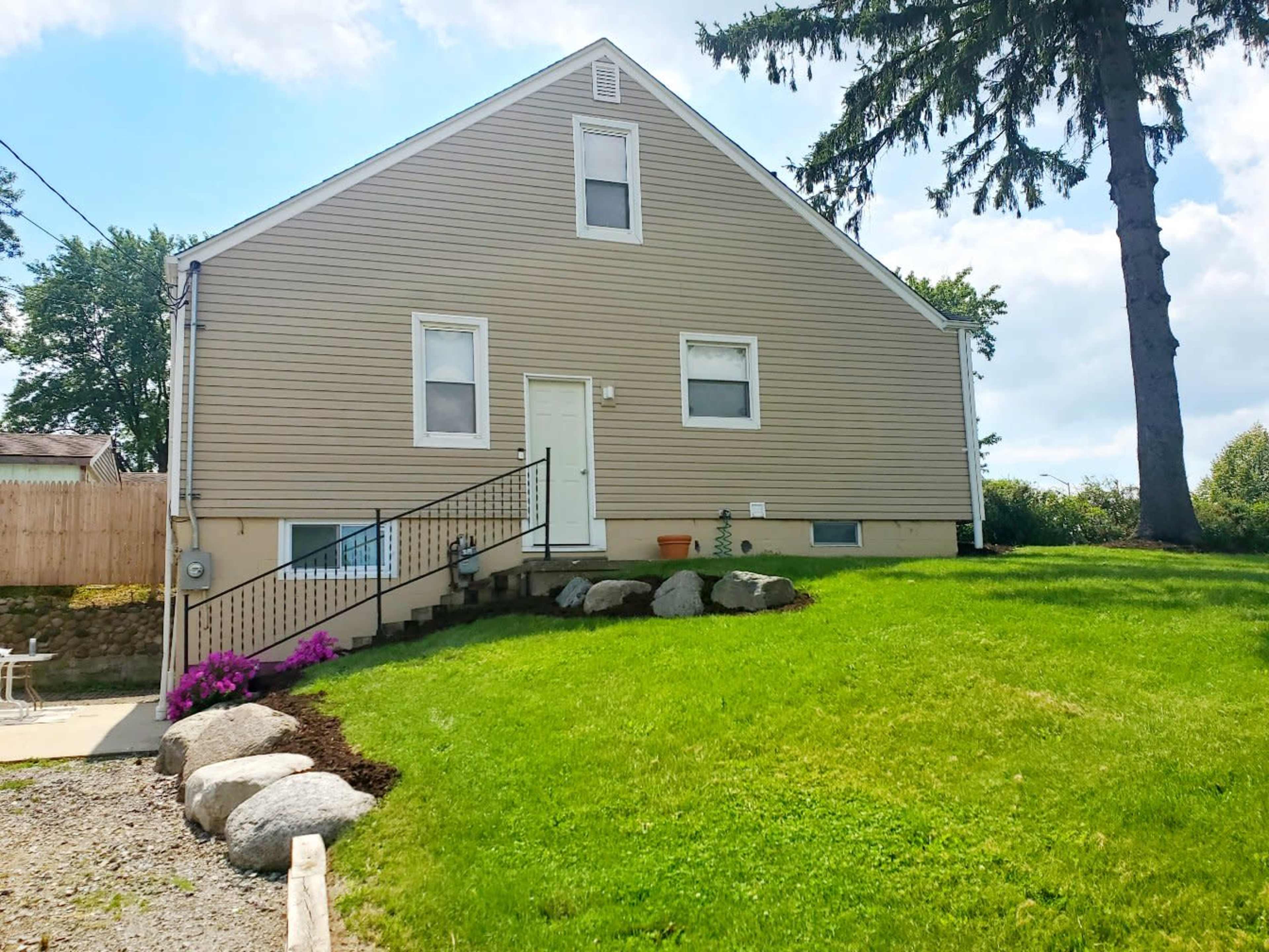 A beige house with a steeply pitched roof sits on a grassy lawn, featuring a set of stairs leading to a front door and bordered by large rocks and flowers.