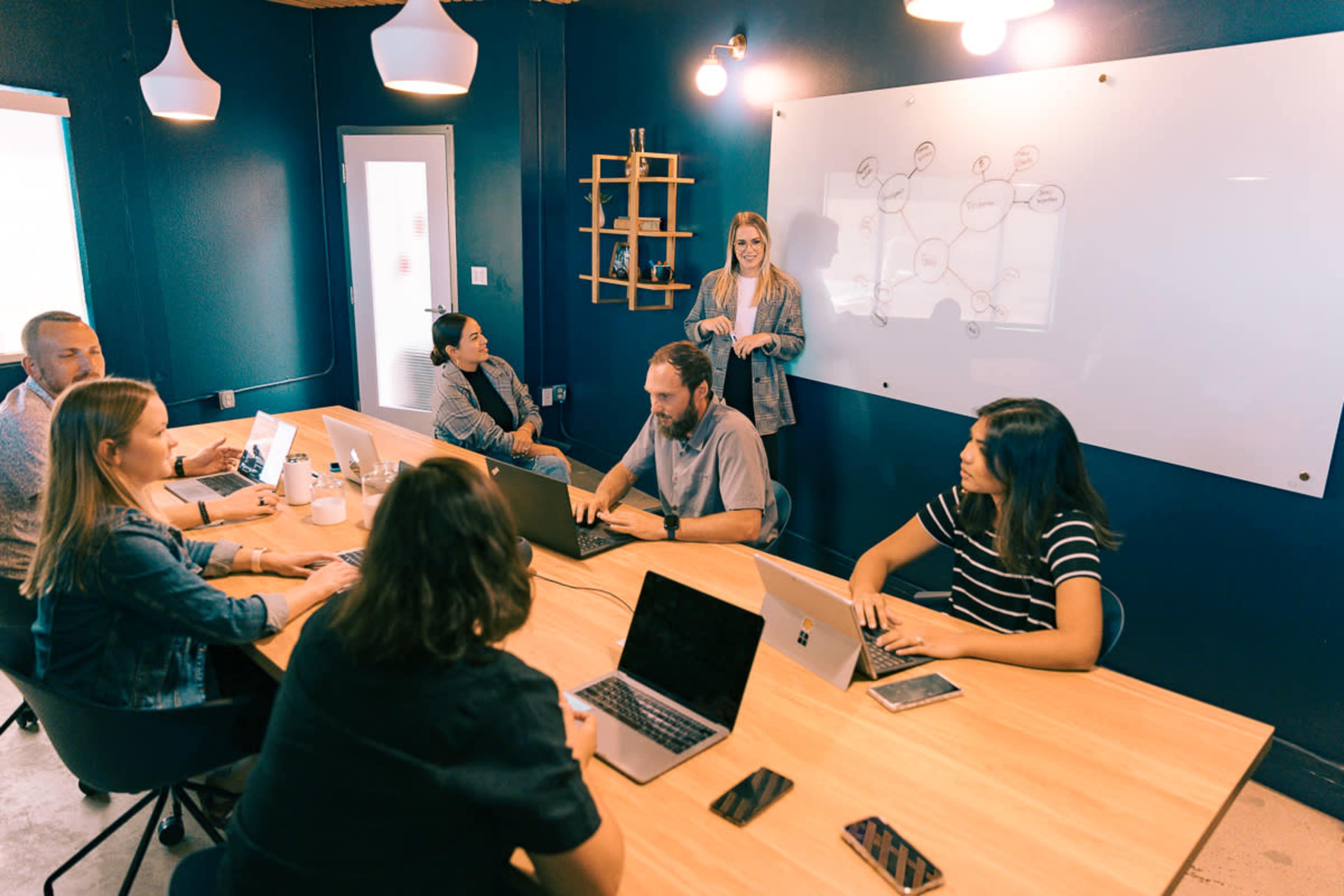 A group of eight people sit around a wooden table in a blue-walled meeting room, with one person standing and presenting in front of a whiteboard.