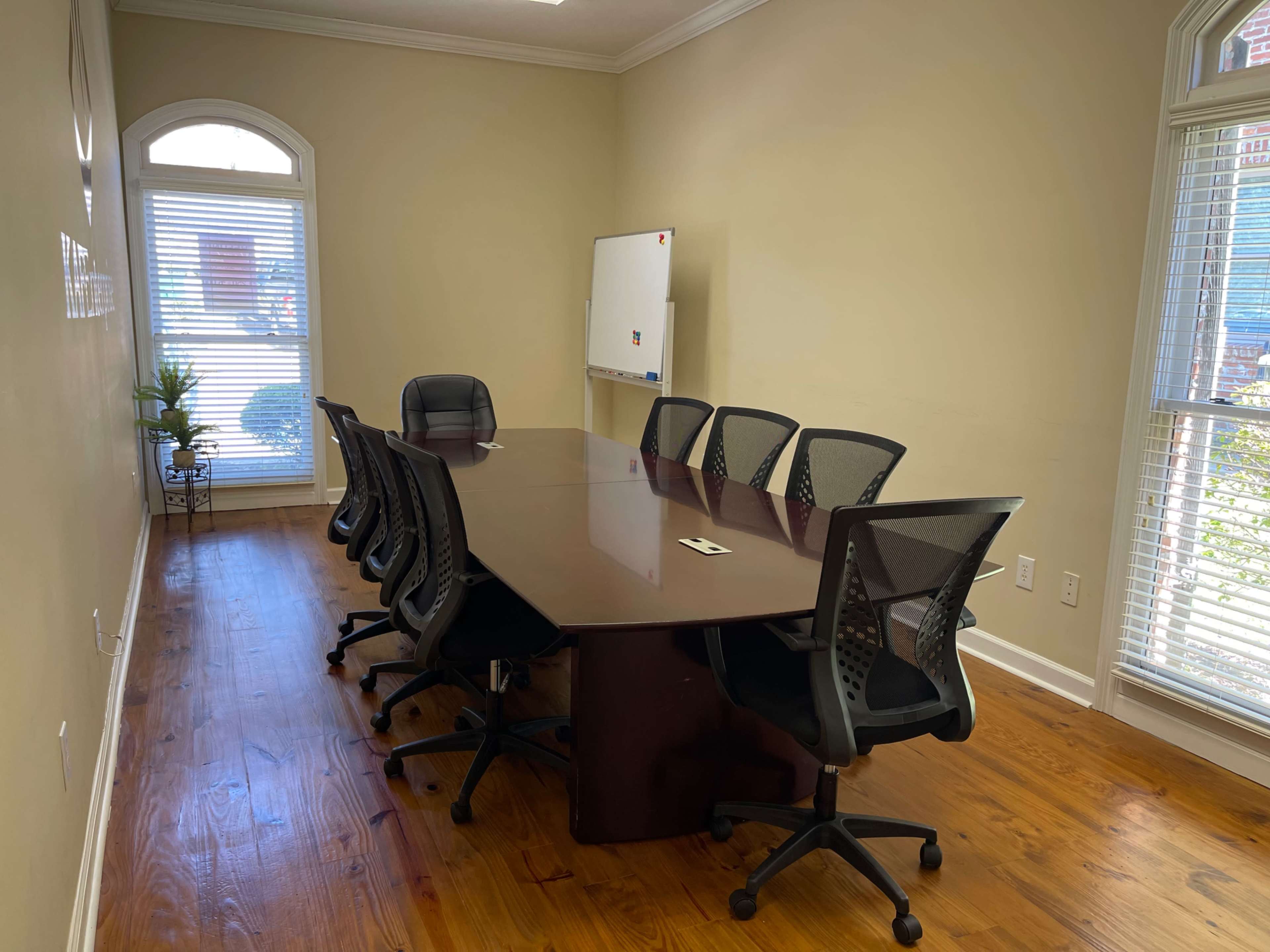 A conference room features a long wooden table surrounded by eight black mesh chairs, with natural light coming through a window and a whiteboard nearby.