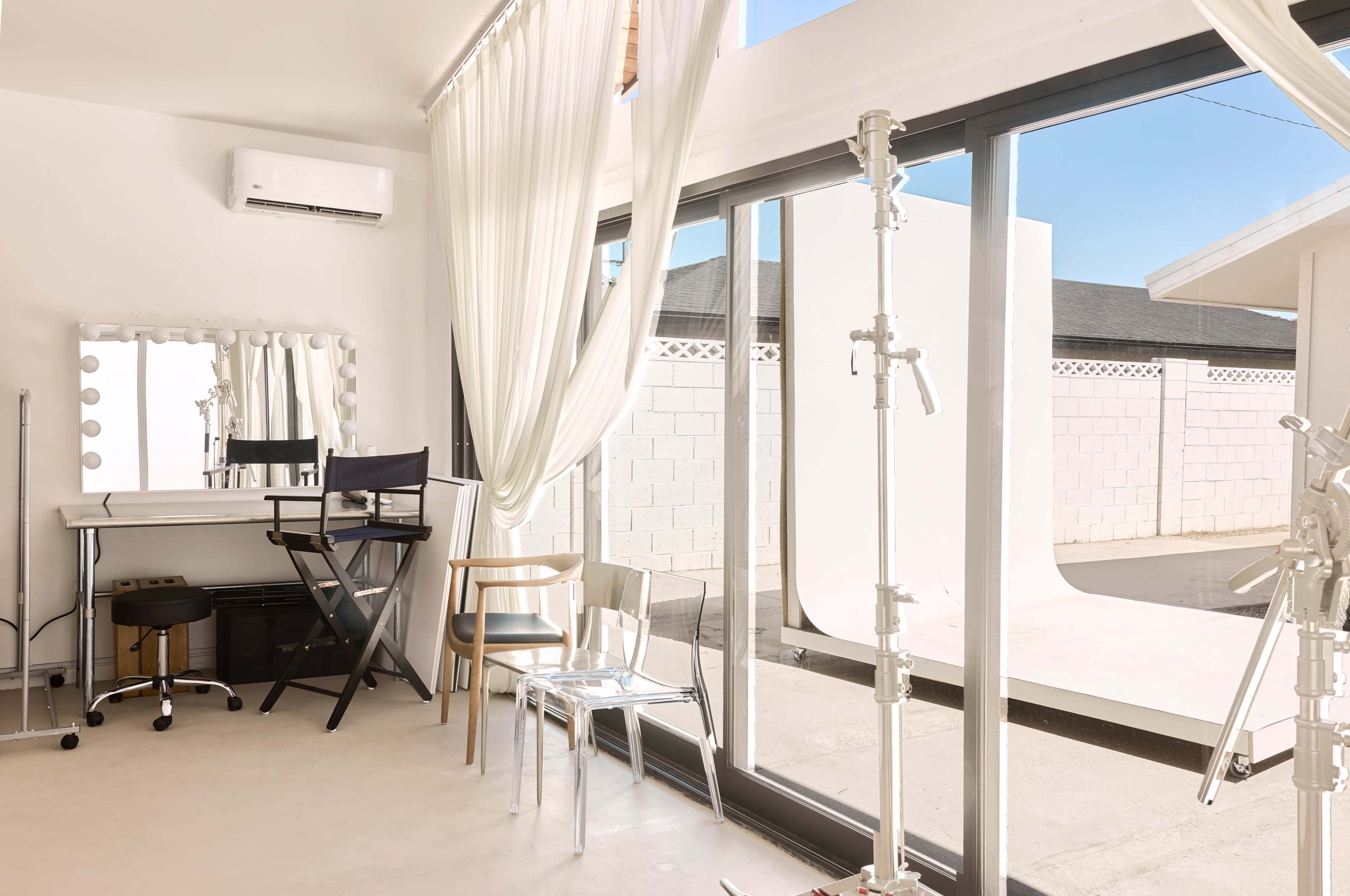 The image shows a modern, bright studio space with large windows, featuring a makeup mirror, chairs, and backdrop support stands.