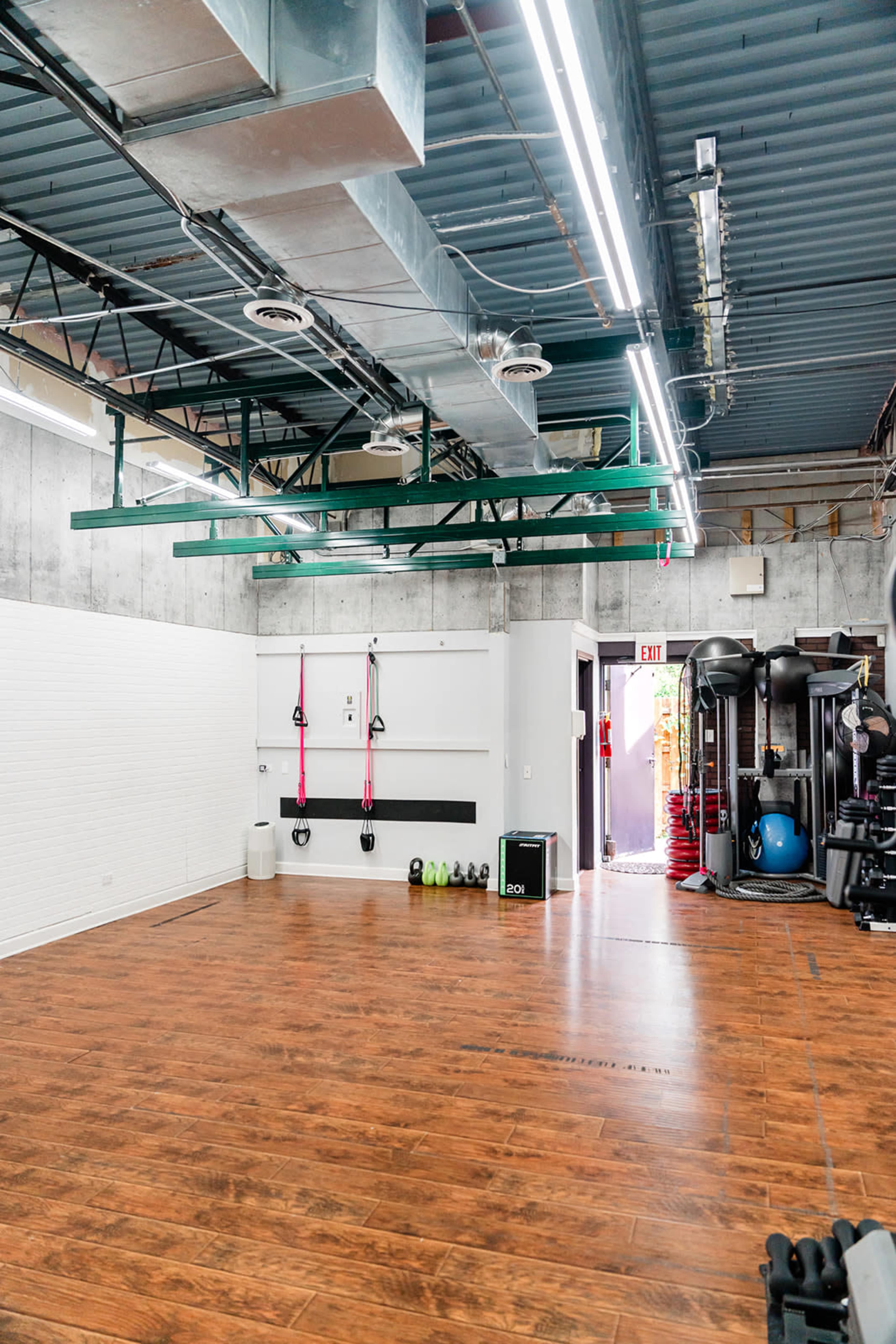 The image shows a spacious gym interior with wood flooring, green overhead equipment racks, and various fitness equipment along the walls.