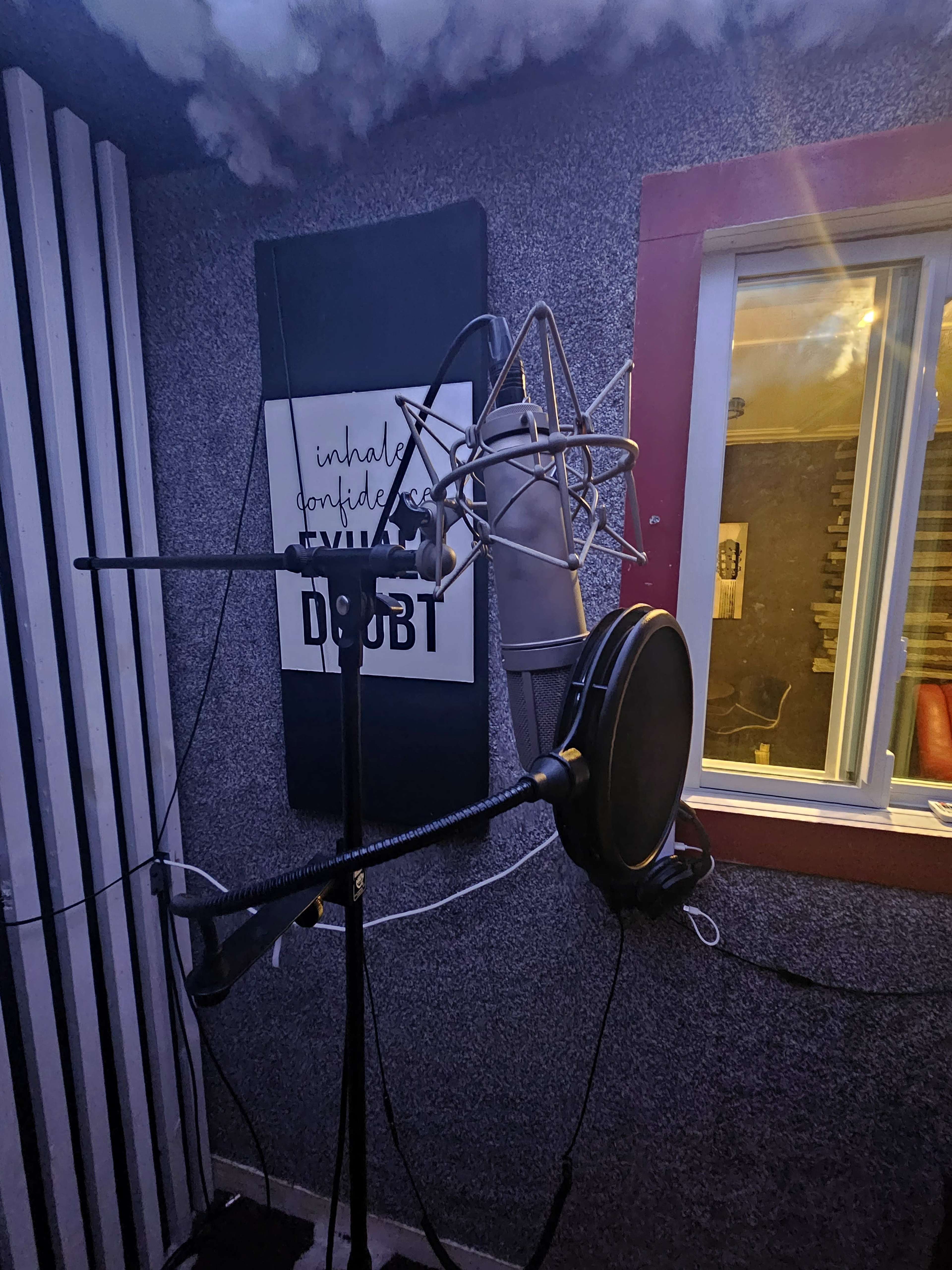 The image shows a microphone setup in a soundproof recording studio with visible acoustic panels and a window in the background.