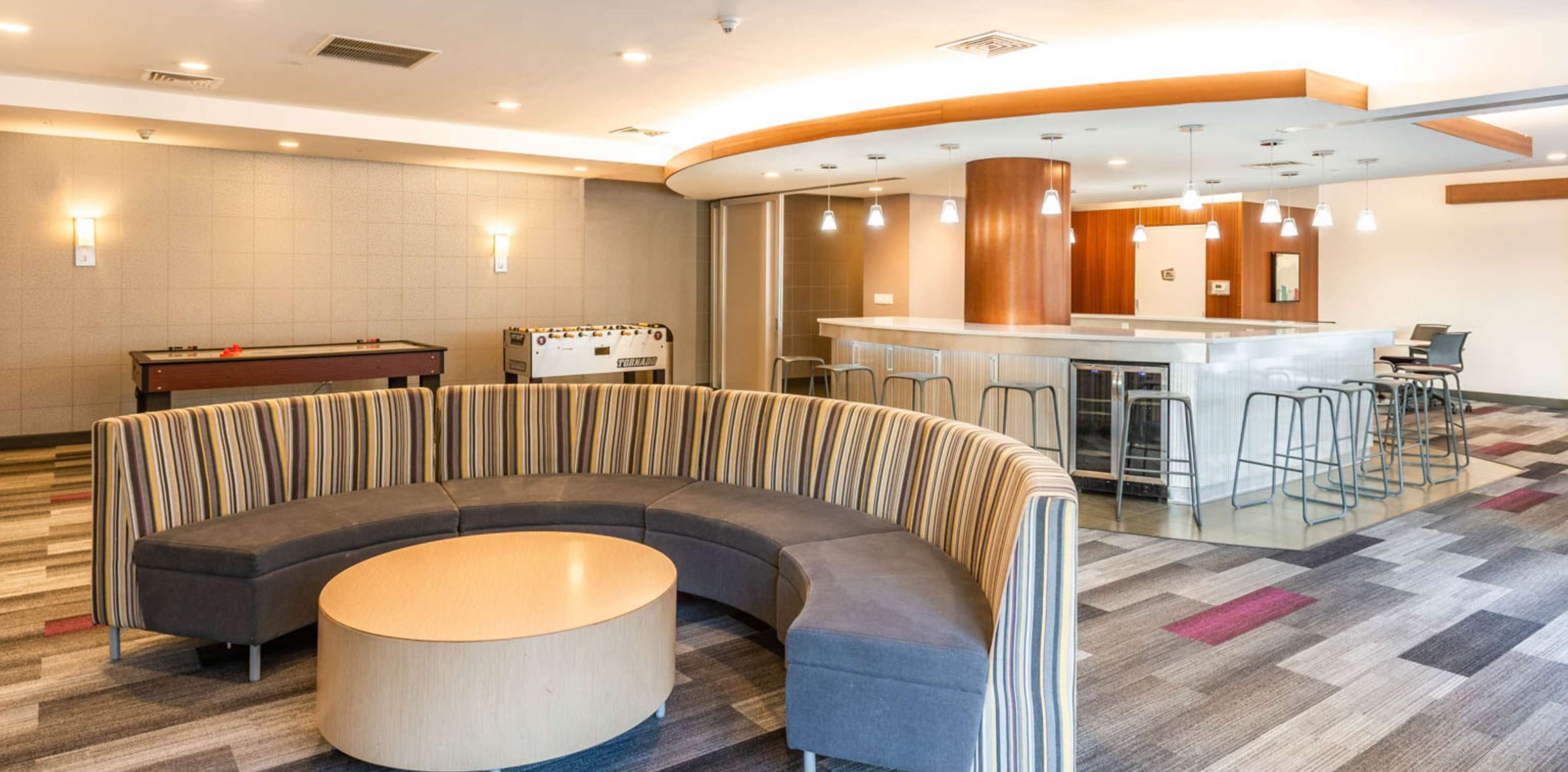 A modern lounge area featuring a curved striped seating arrangement with a circular wooden table, a game table, and a bar counter with stools in a spacious room.