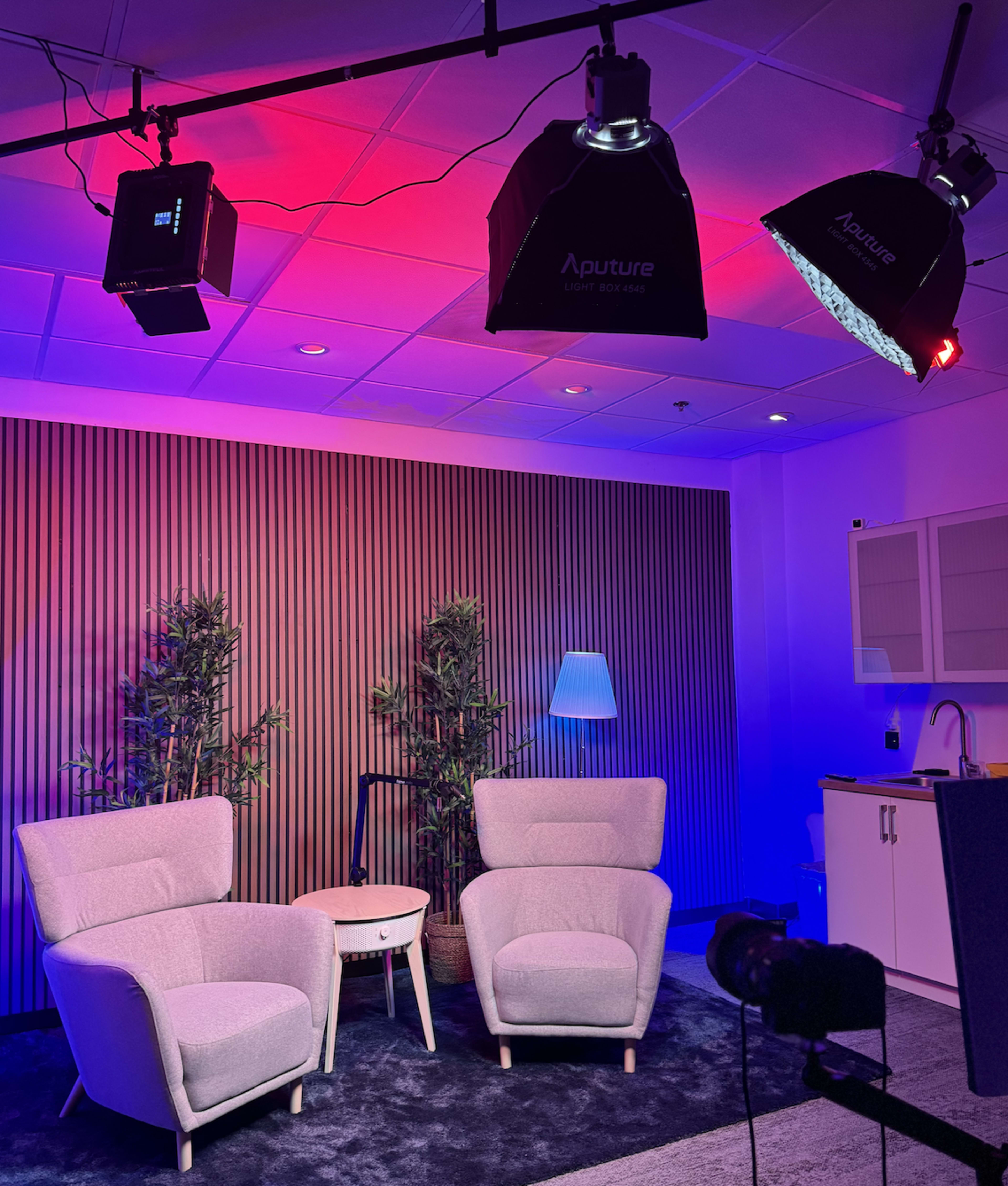 A modern studio setup with two light-colored armchairs, a small side table, and soft lighting against a background of wooden slats and plants.