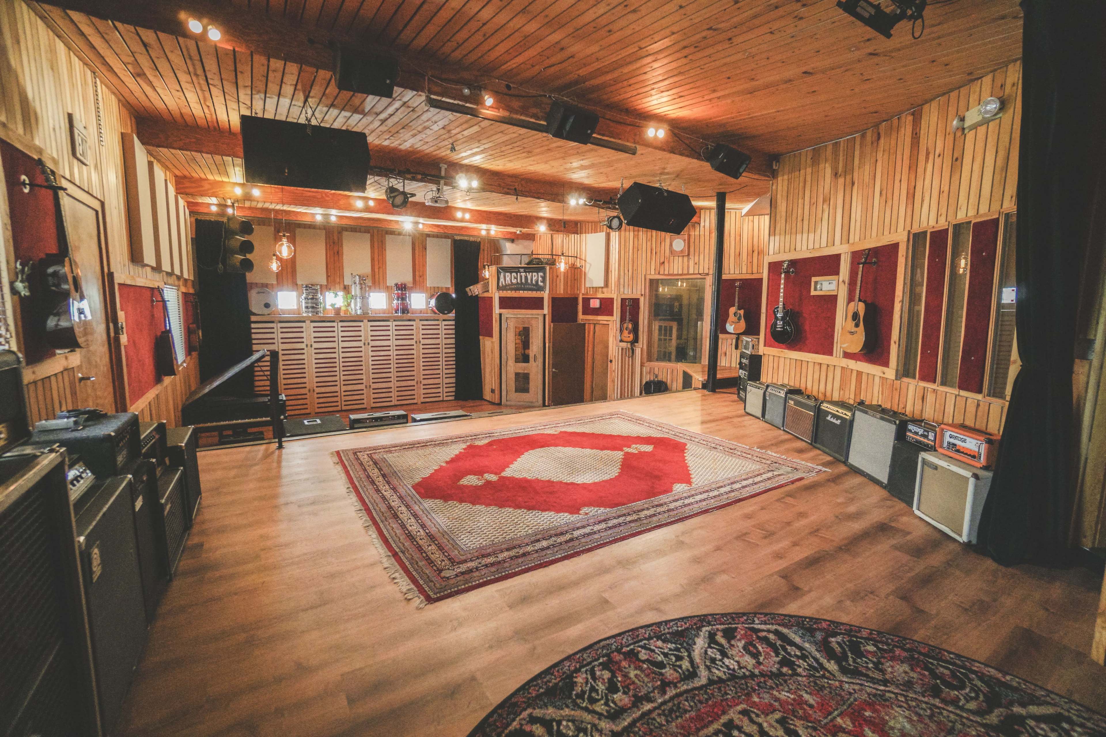 The image shows a wooden-paneled recording studio with amplifiers and instruments arranged along the walls and a large rug on the floor.