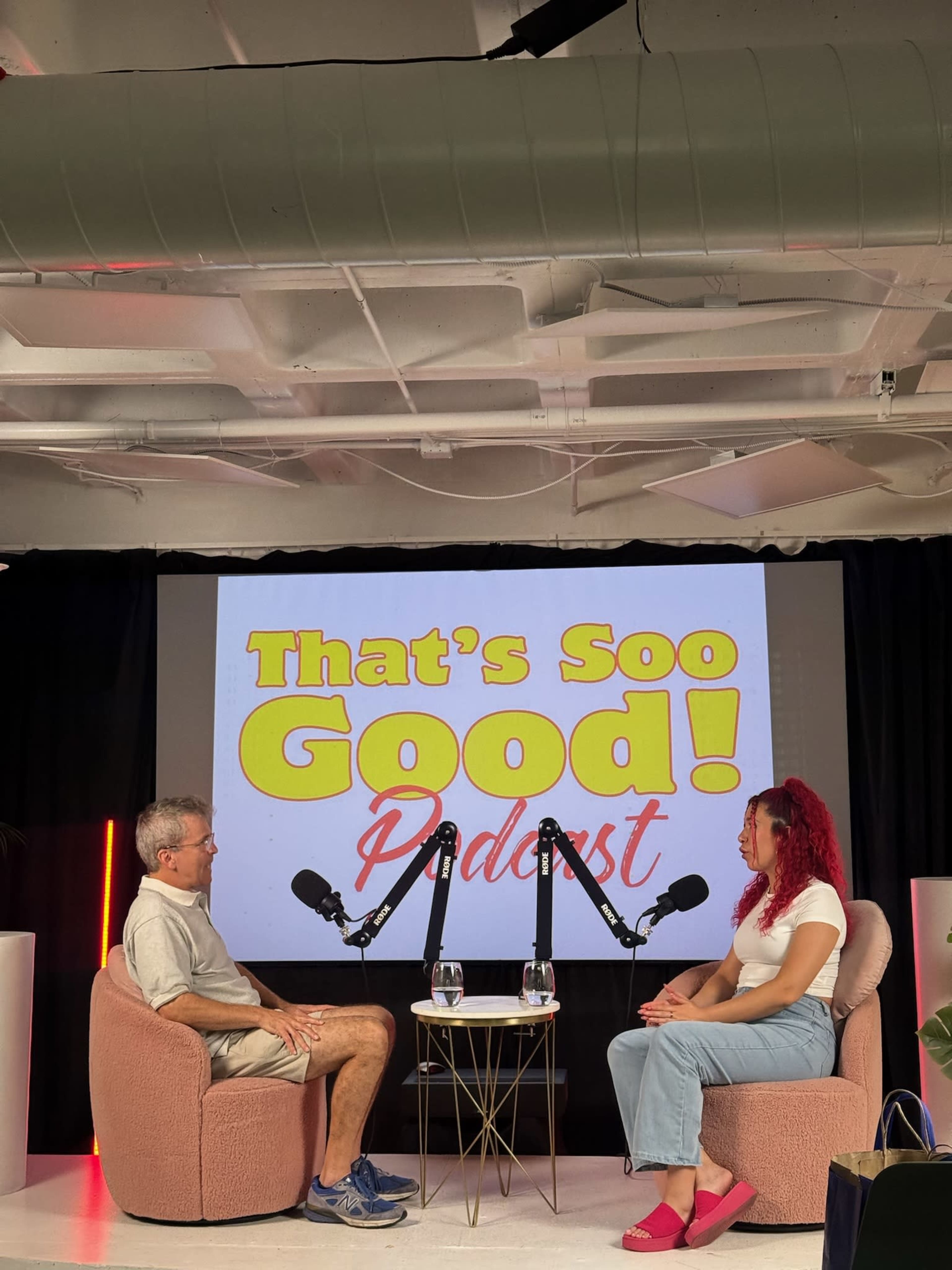 A man and a woman are seated on chairs in a podcast studio with a backdrop displaying the title "That's Soo Good! Podcast" and microphones in front of them.