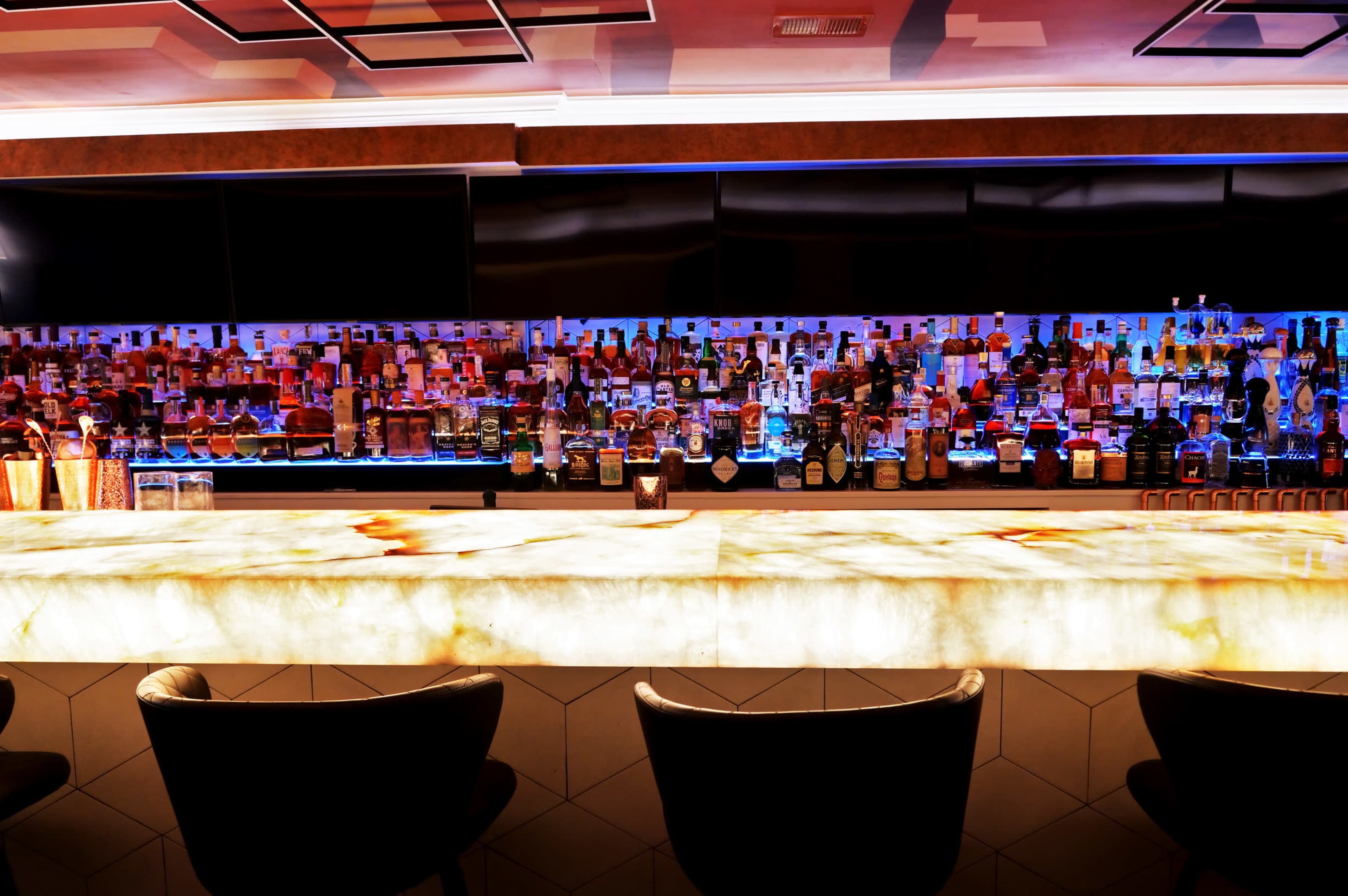 The image shows a modern bar with a brightly lit, illuminated countertop and shelves lined with various bottles of alcohol in the background.