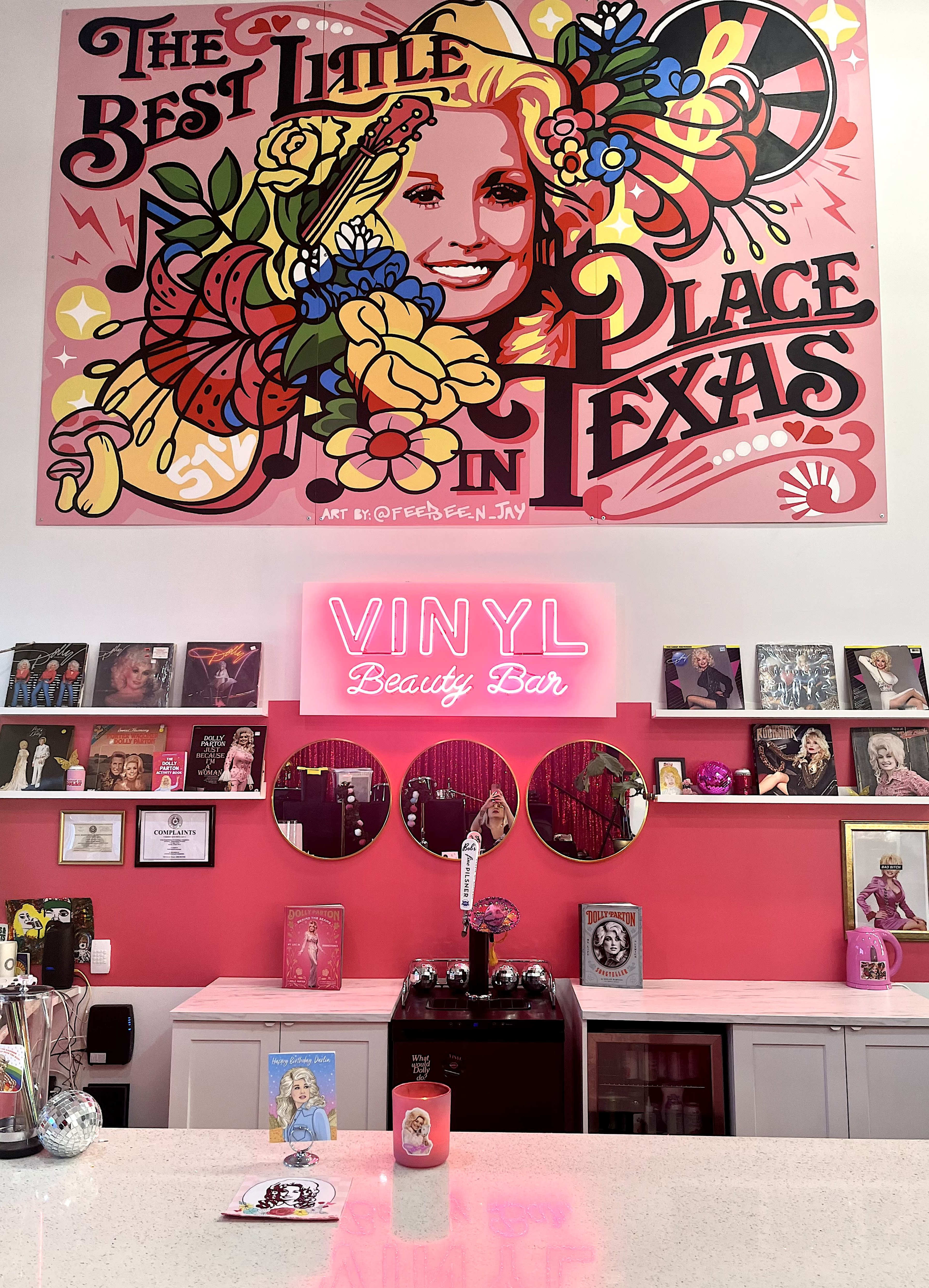 The image shows a brightly colored beauty bar with a vibrant mural above, featuring floral designs and the text "The Best Little Place in Texas," alongside a neon sign that reads "VINYL Beauty Bar."
