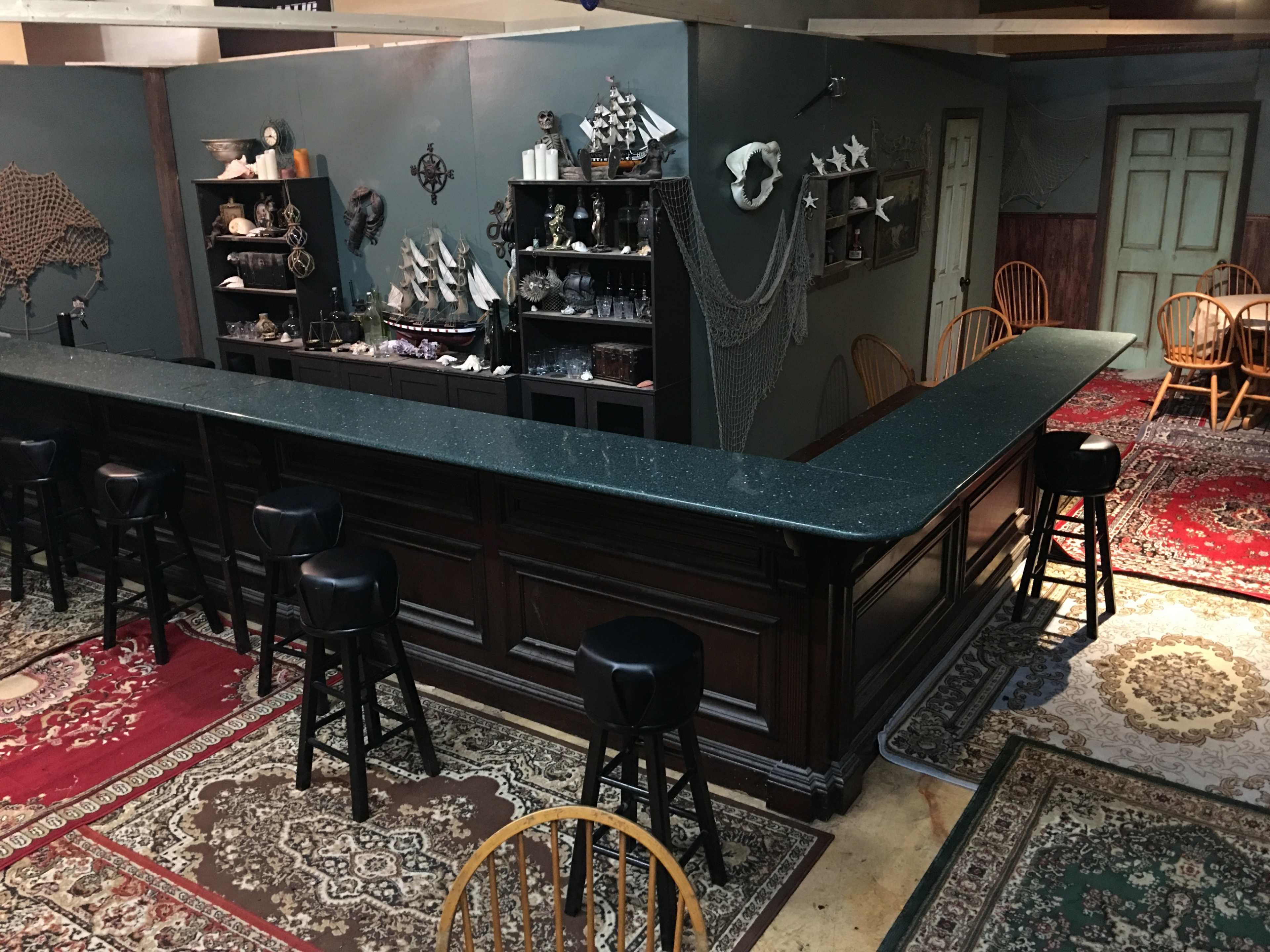 A dimly lit bar area with a long wooden counter, black stools, and various decorative items displayed on shelves against dark walls, all set on patterned carpets.