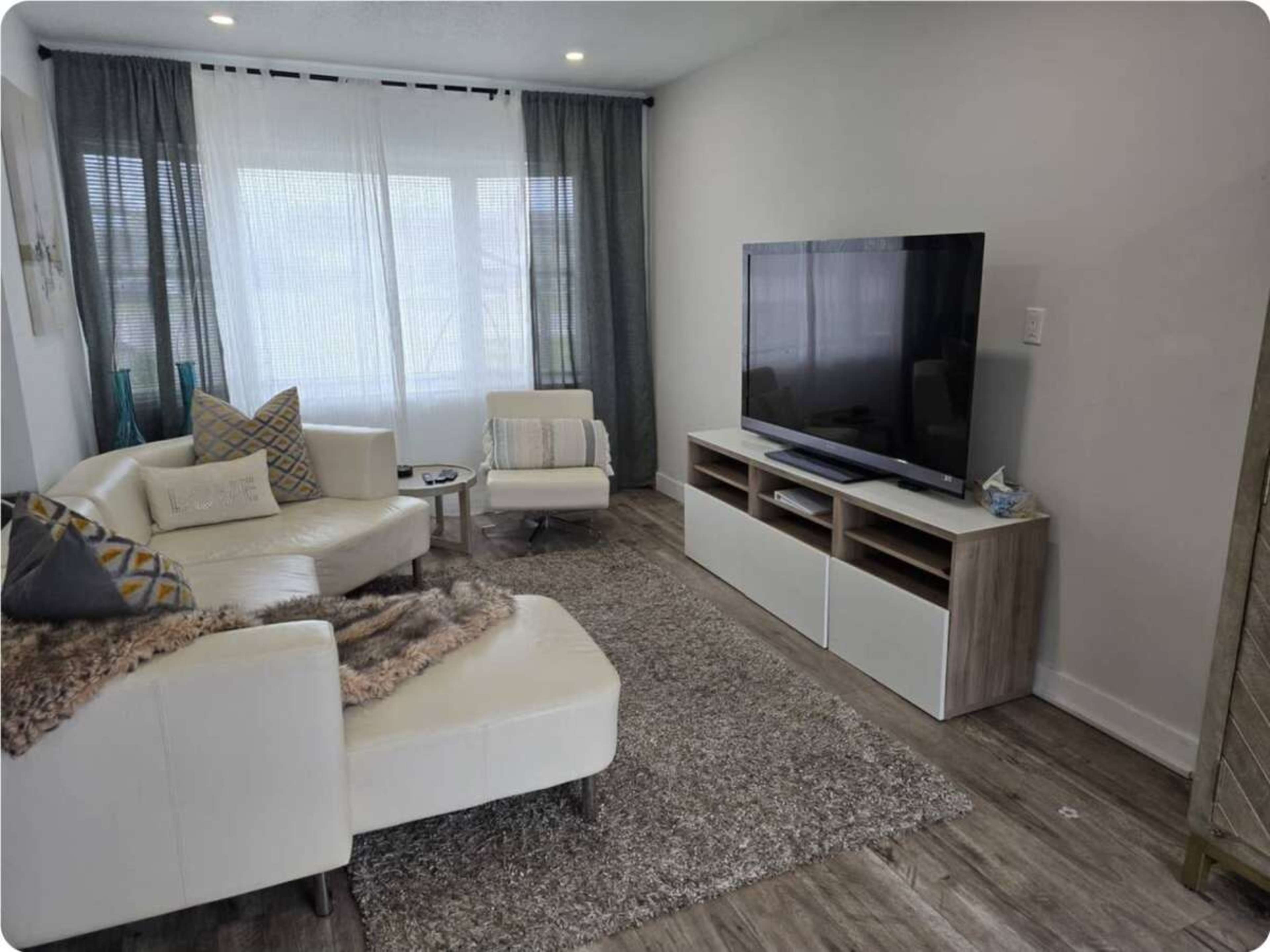 The image shows a modern living room with a white sectional sofa, a small armchair, and a television mounted on a light wood media console.