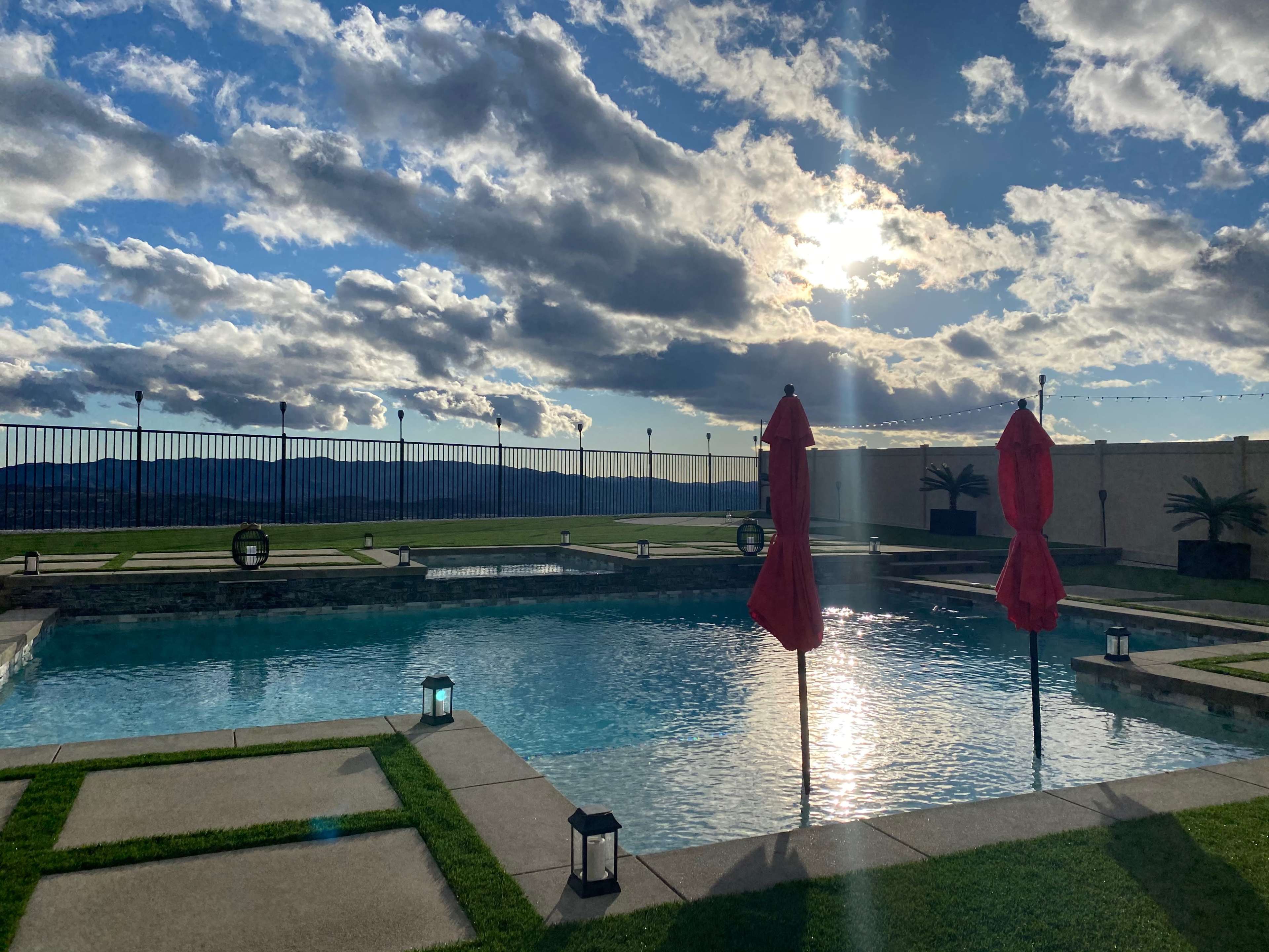 The scene features a swimming pool with two red umbrellas beside it, set against a backdrop of clouds and a setting sun.