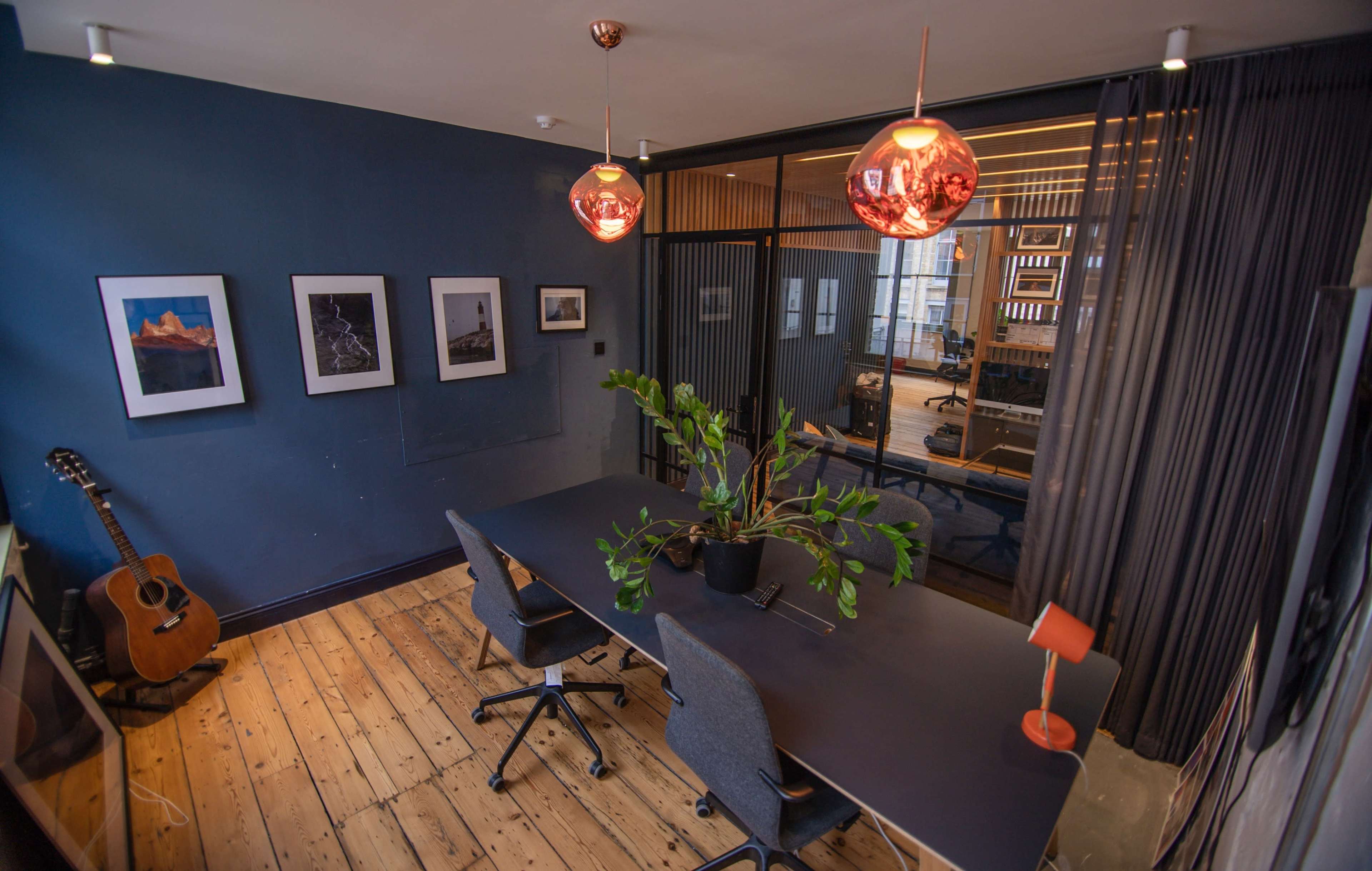 A modern office space featuring a long table with four chairs, a potted plant in the center, wall art, and two pendant lights, with a guitar resting against the wall.