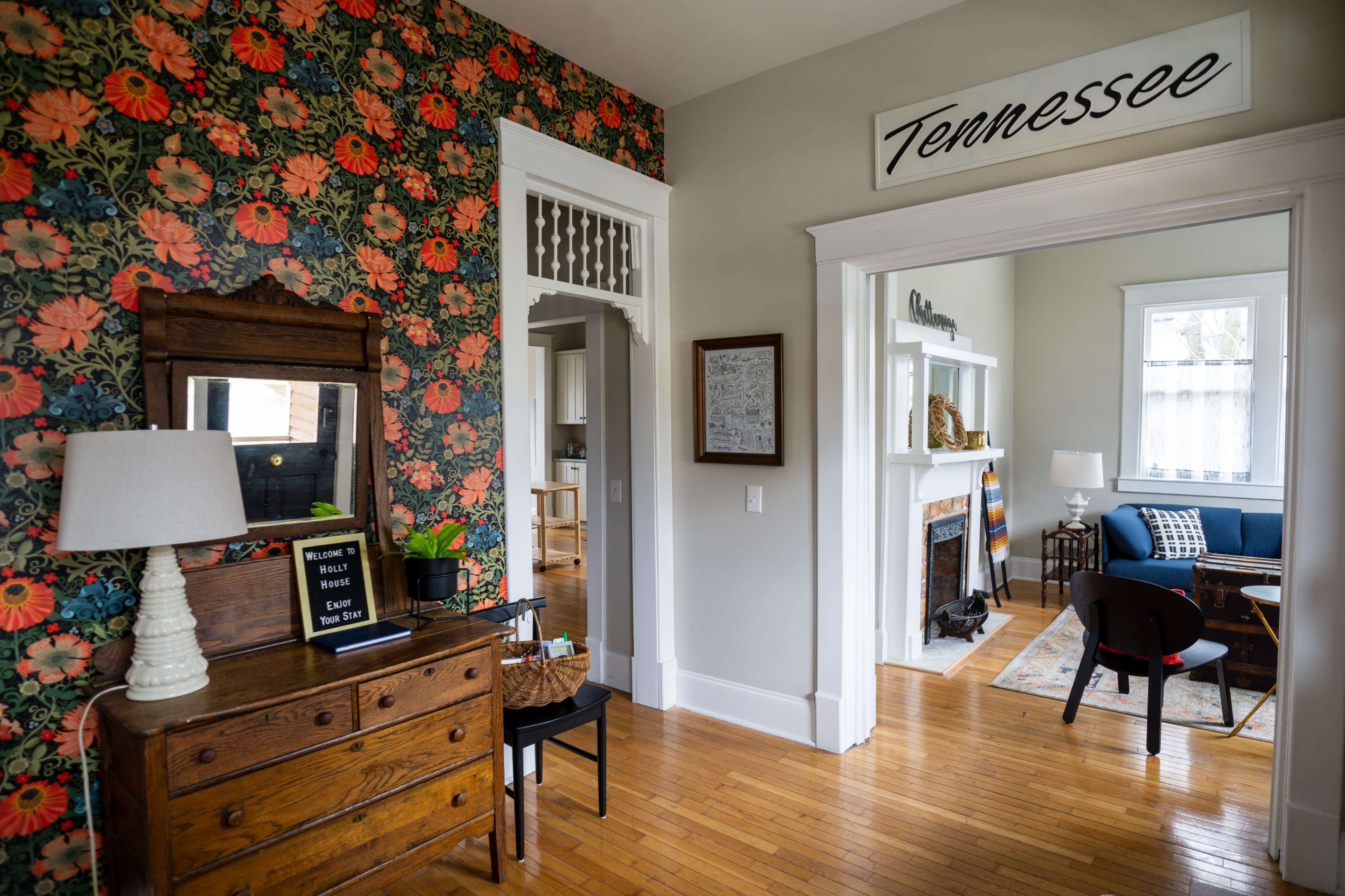 The image shows a living space with floral wallpaper, a wooden dresser with a lamp, and a cozy seating area visible through an open doorway.
