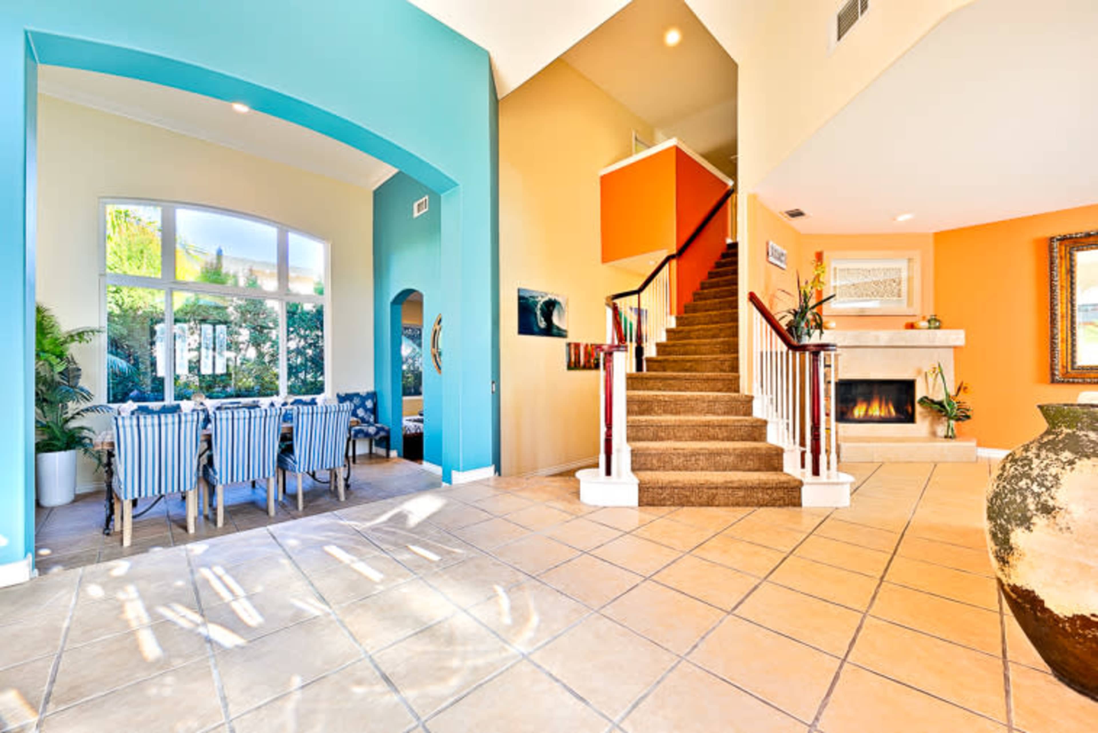 A spacious living area featuring a staircase, a dining area with striped chairs, and large windows allowing natural light to enter.