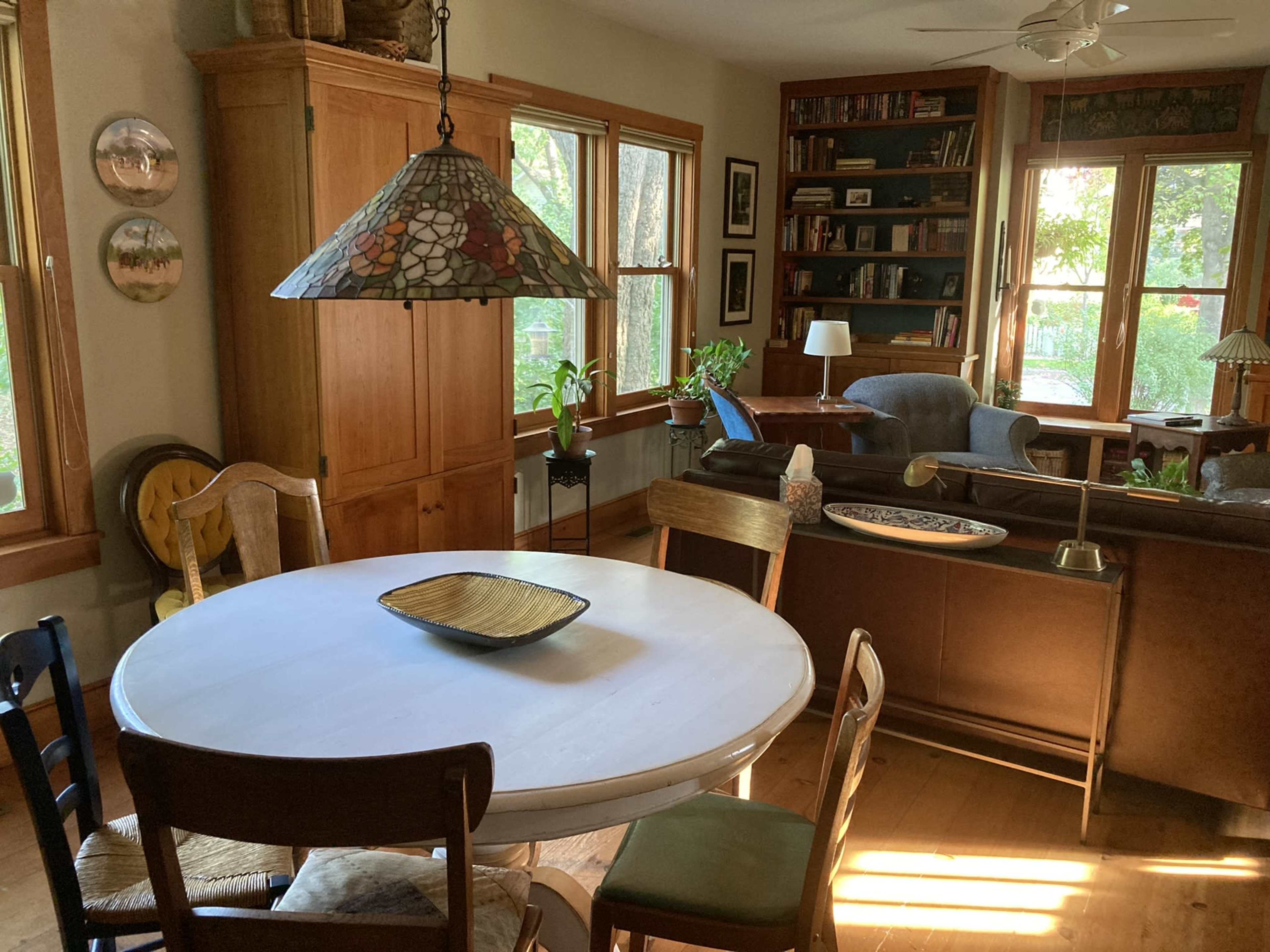 A dining area with a round white table surrounded by chairs, leading into a living room filled with bookshelves and large windows.