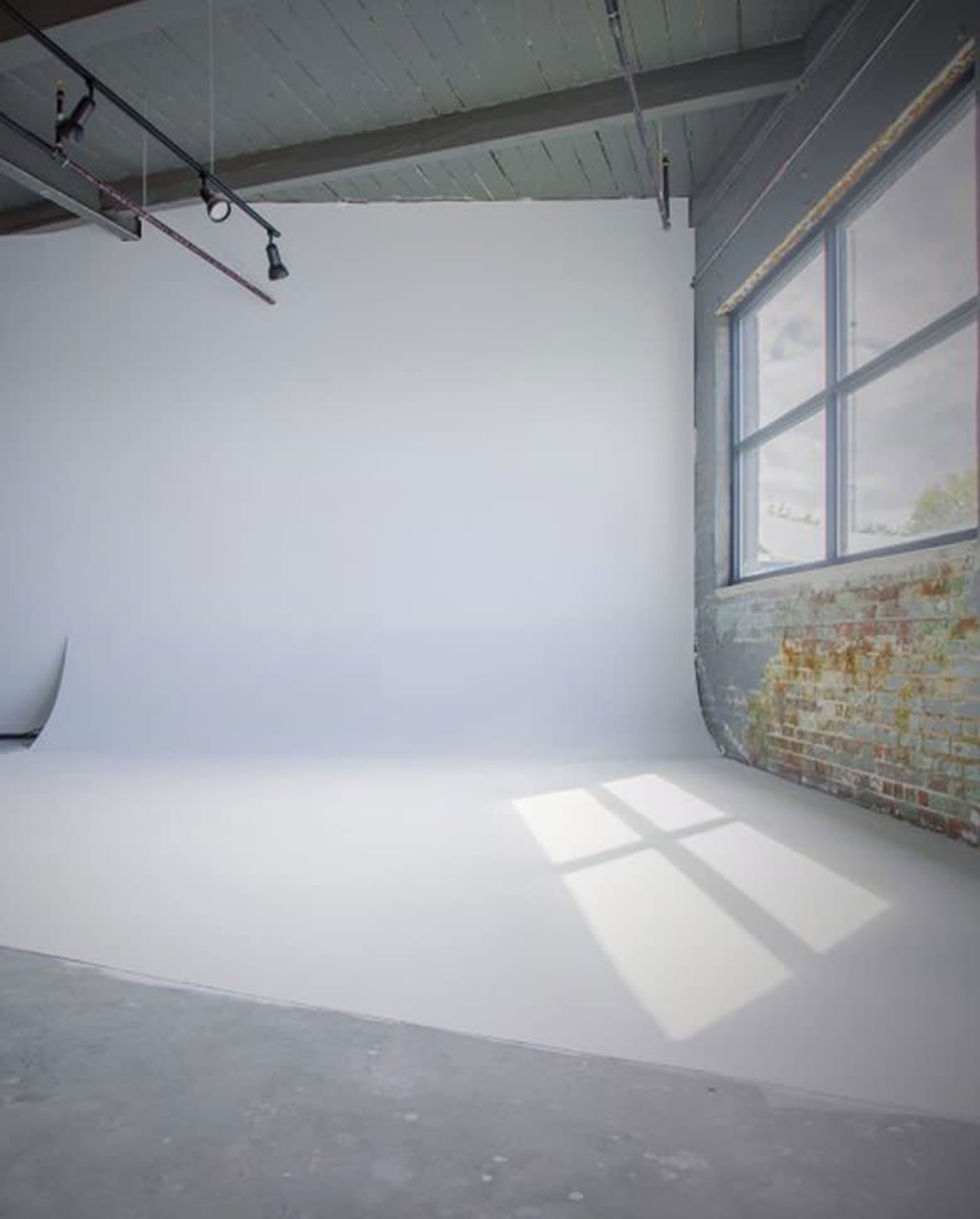 The image shows a spacious photography studio with a white backdrop, large window, and exposed brick wall.