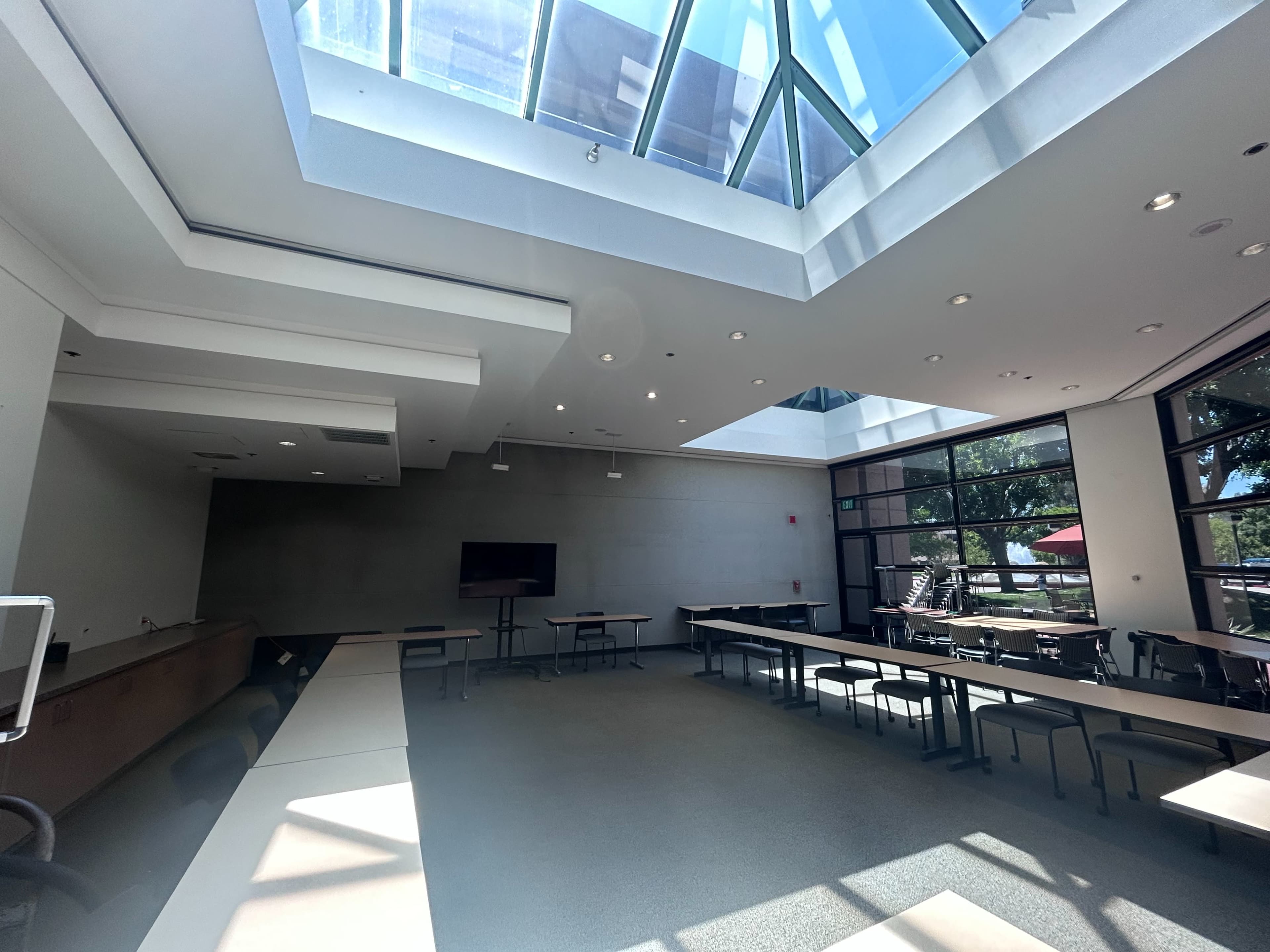 A spacious conference room with large windows and a skylight, featuring several long tables and a television screen at one end.