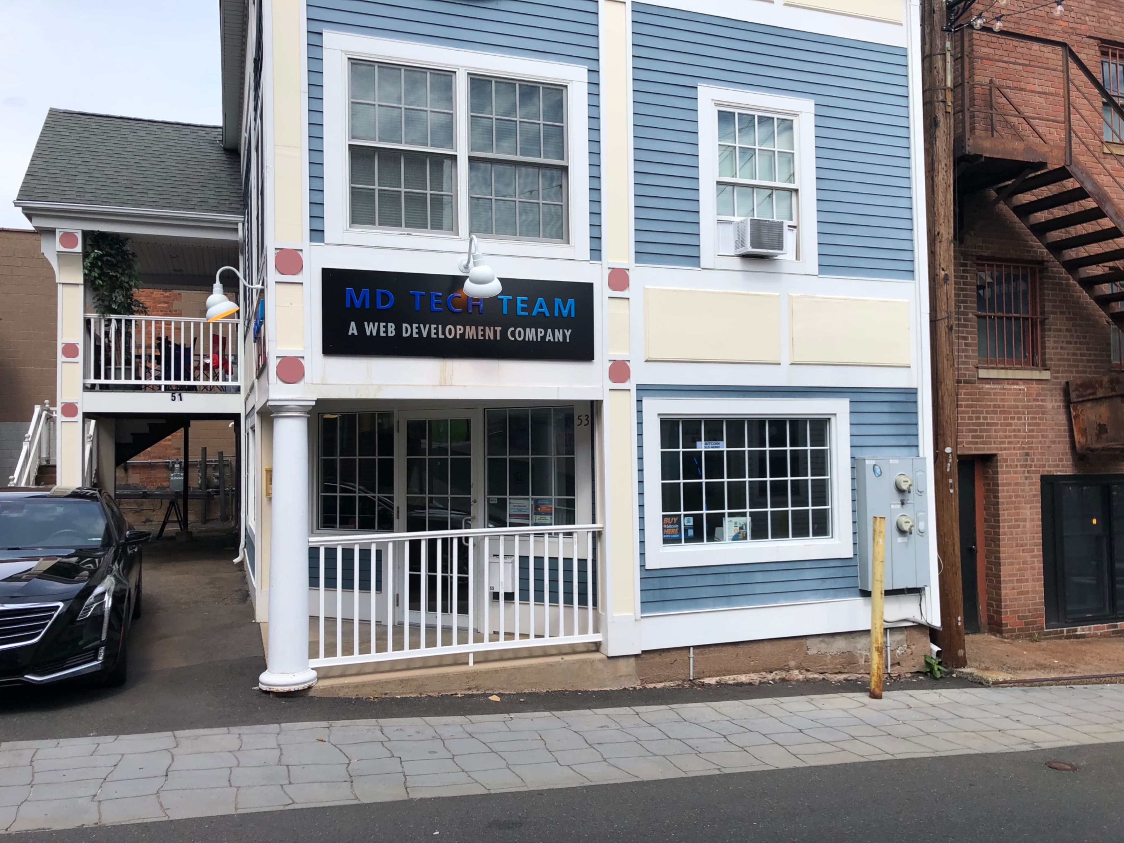A multi-story building with blue and white paneling houses a sign for "MD Tech Team," a web development company, next to an adjacent parking area.