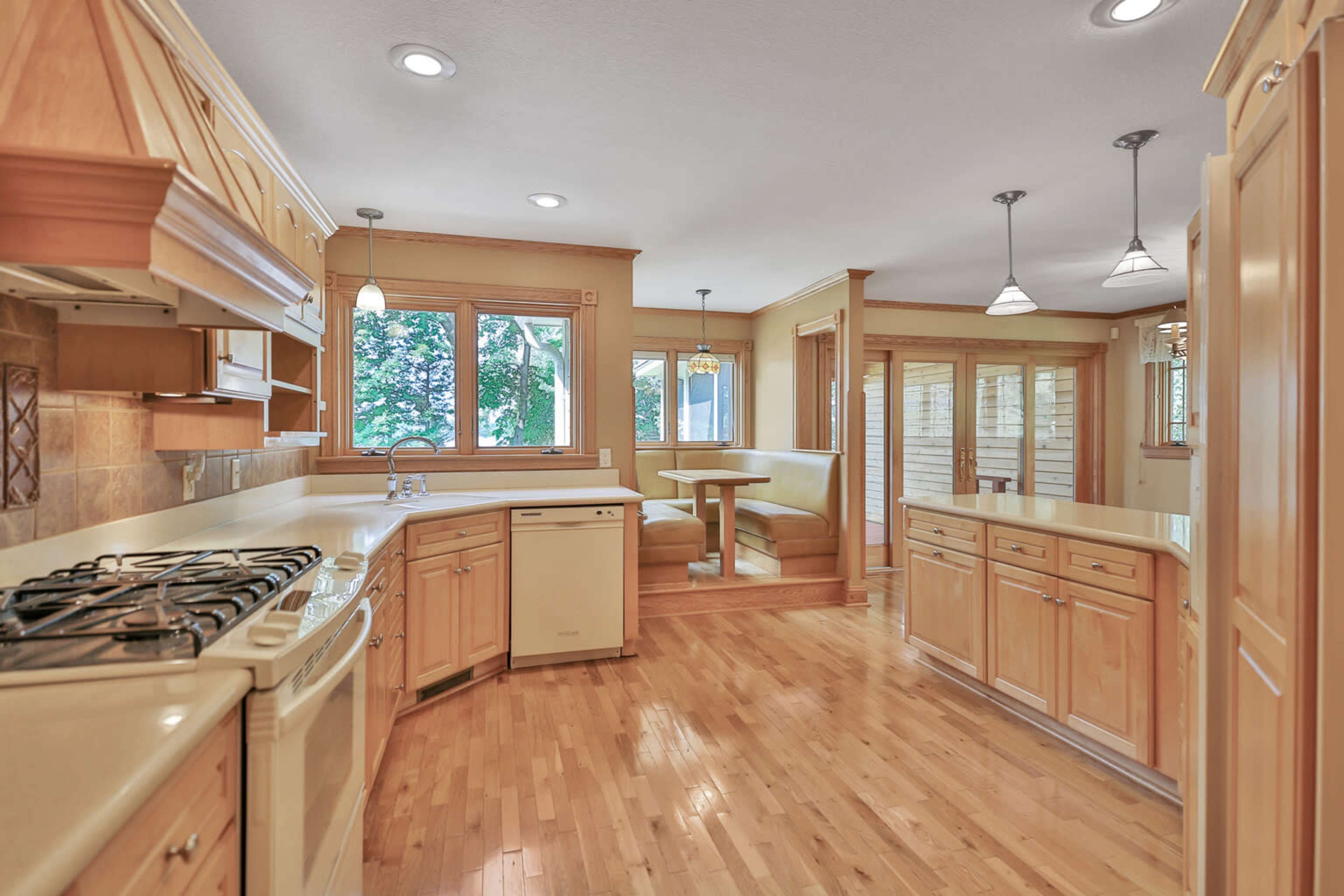 The image shows a well-lit kitchen featuring wooden cabinetry, a gas stove, and a small dining area with a circular table and benches.