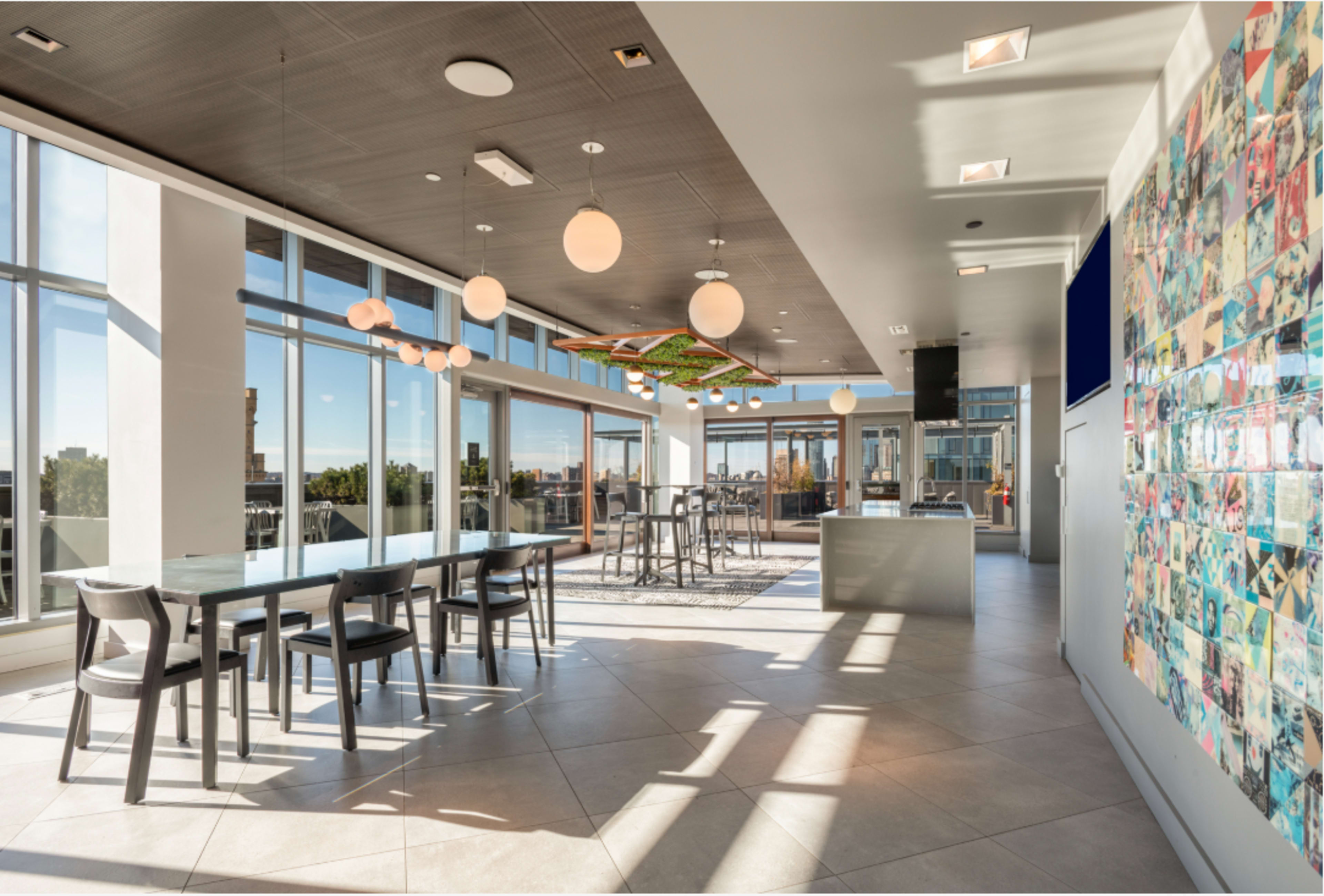 The image depicts a modern open-concept kitchen and dining area featuring large windows, a long table with black chairs, and a colorful wall display.
