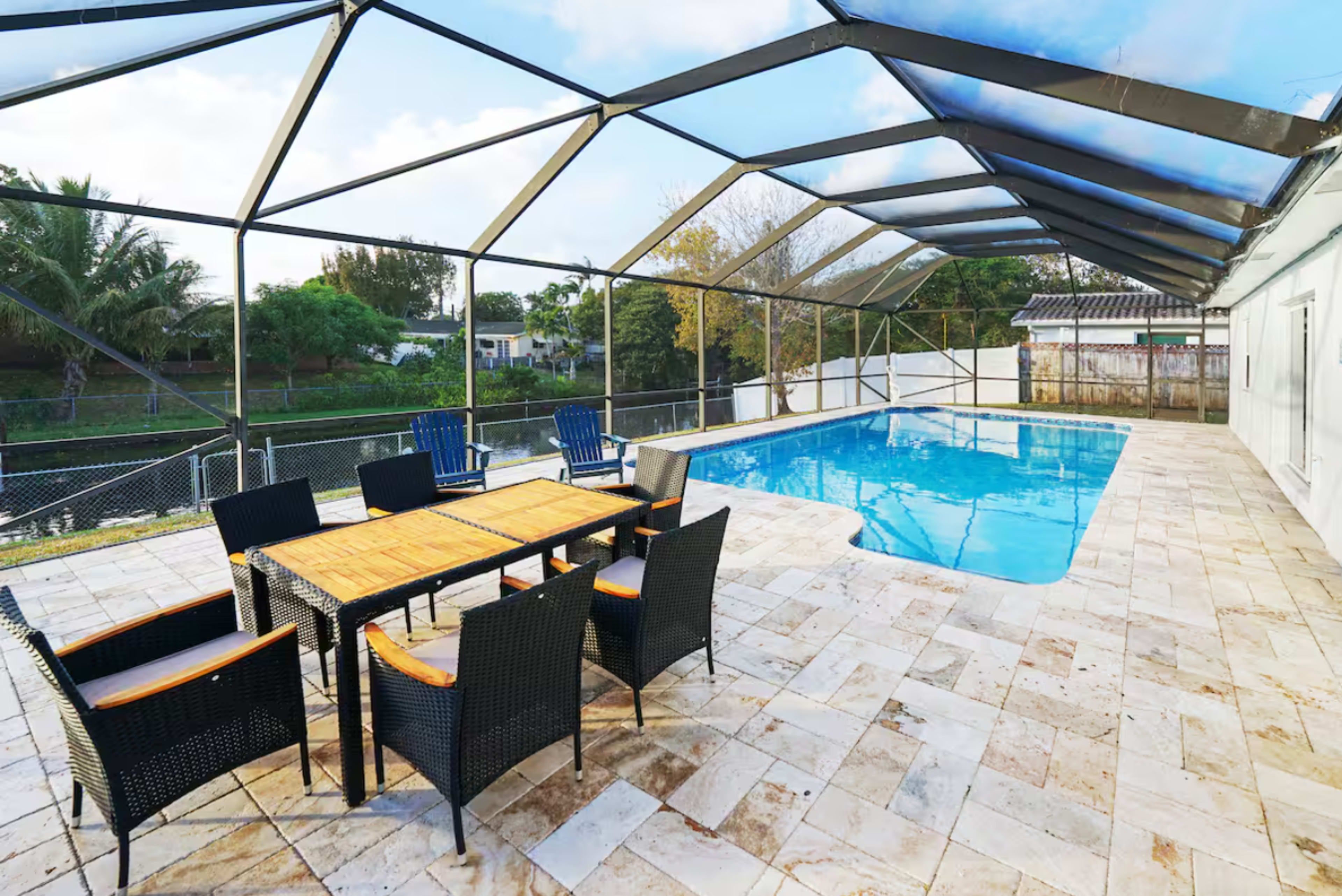 A fenced outdoor patio features a swimming pool surrounded by a table and chairs under a partially covered roof.