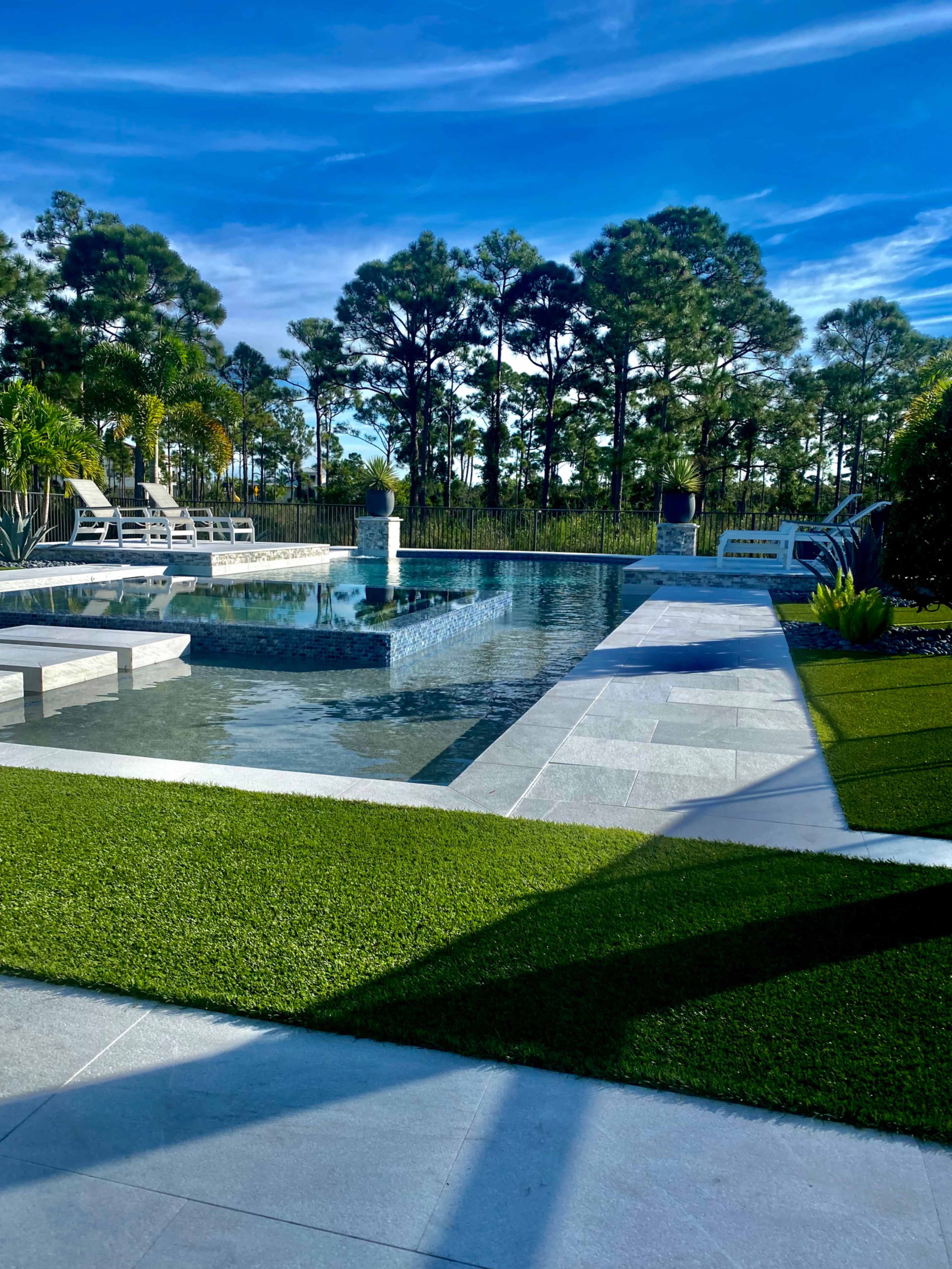 A modern outdoor swimming pool area surrounded by green grass and trees under a blue sky.