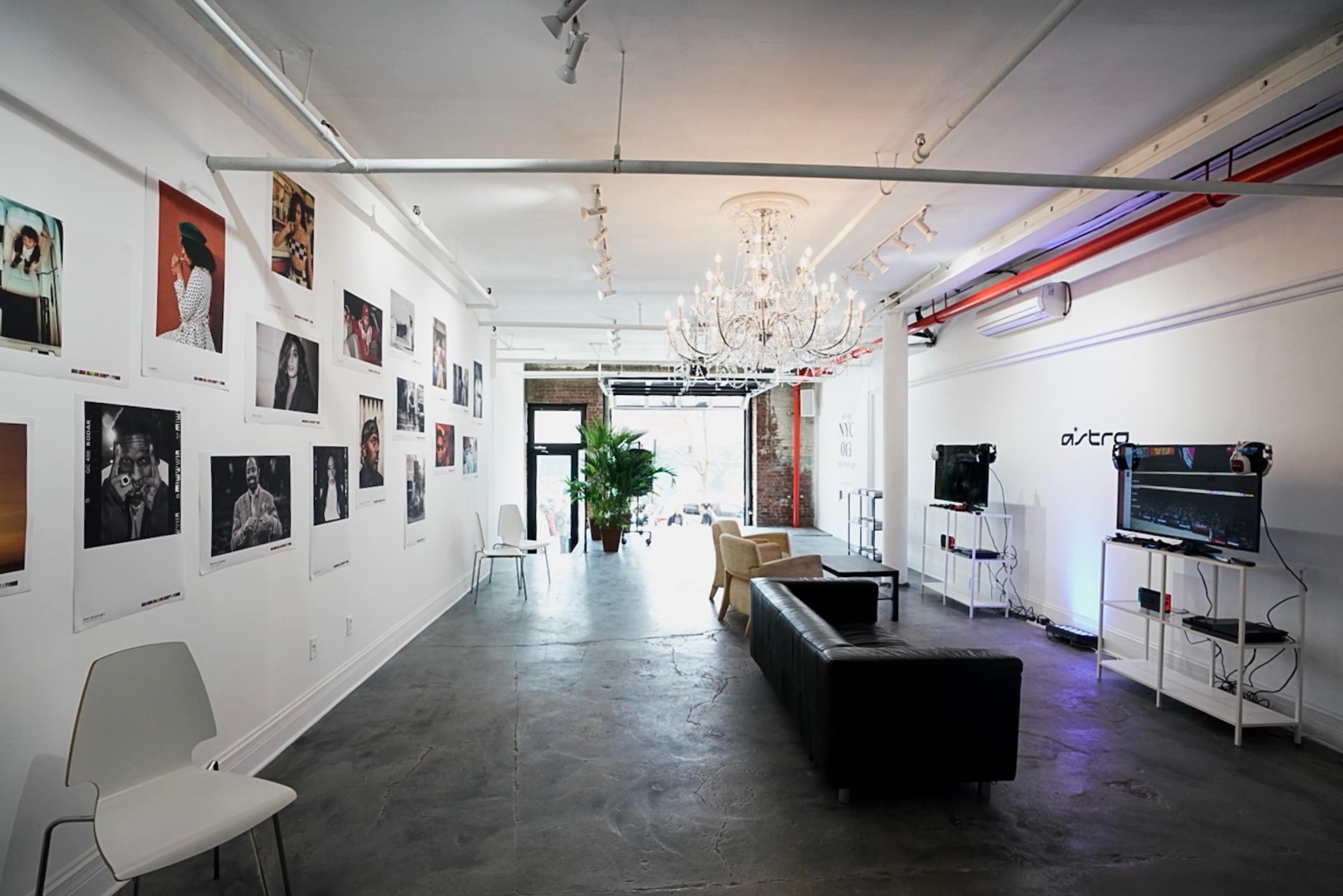A modern art gallery with exposed brick walls, framed photographs displayed on the white walls, and a seating area featuring a black sofa and a light-colored chair.