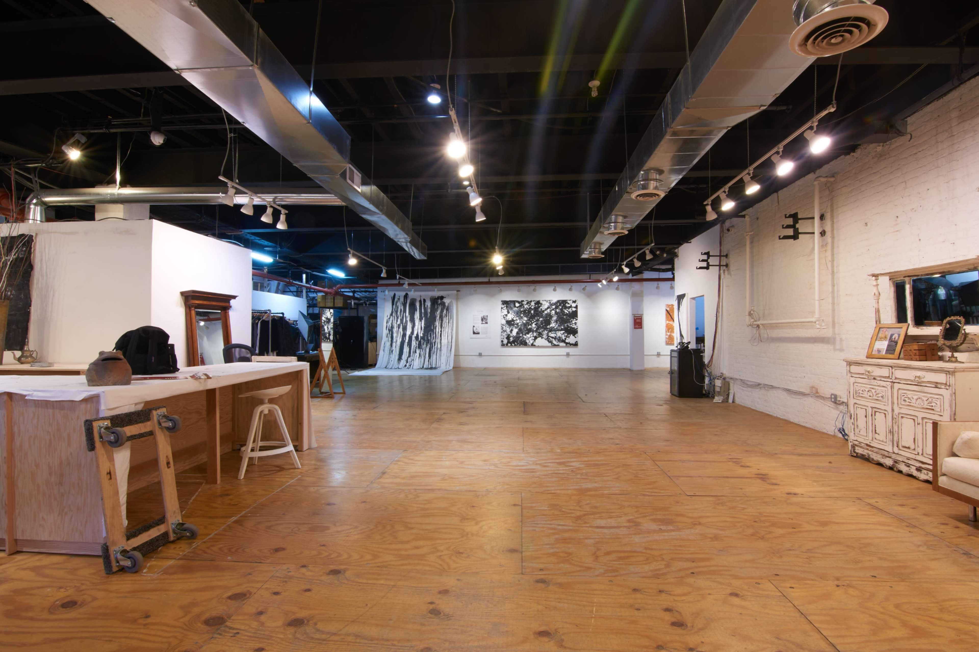 The image shows a spacious art gallery with wooden floors, white walls displaying black and white artwork, and various art supplies on a worktable.