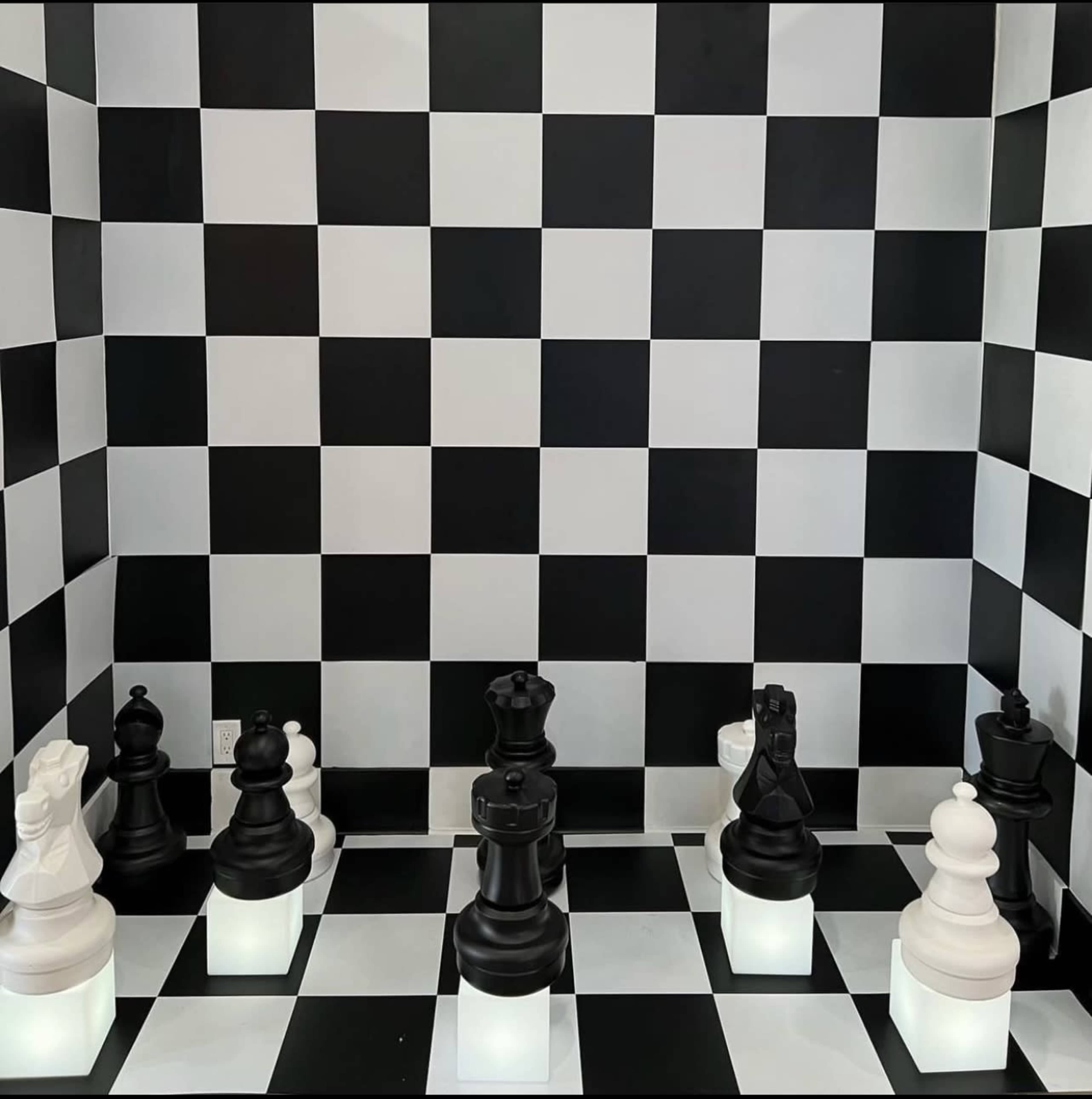 A chess game is set up on a black and white checkered board, featuring large black and white pieces illuminated from below.