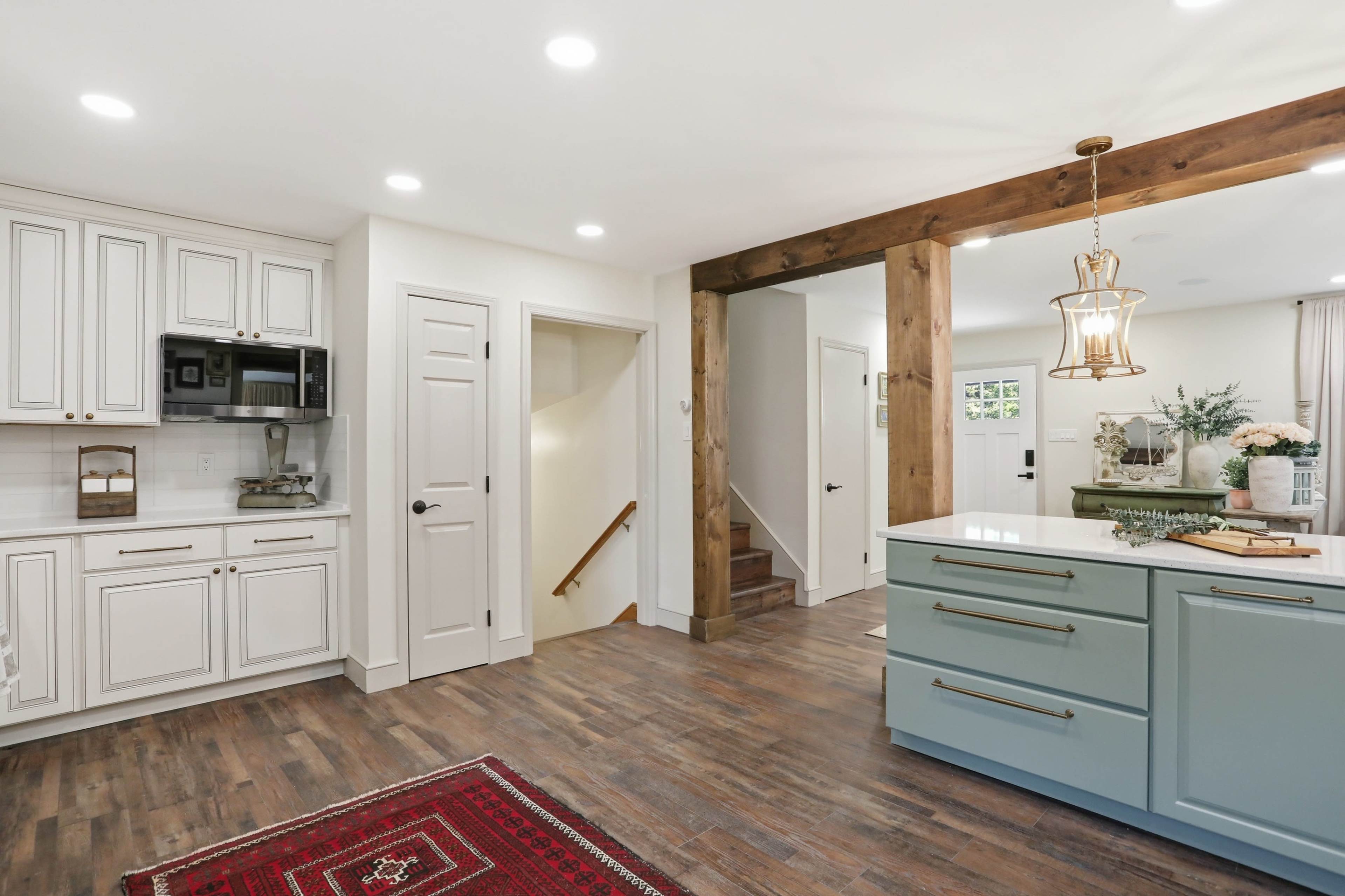 The image shows a modern kitchen with white cabinets, a light blue island, and wooden accents, connected to a hallway with stairs and a front door.
