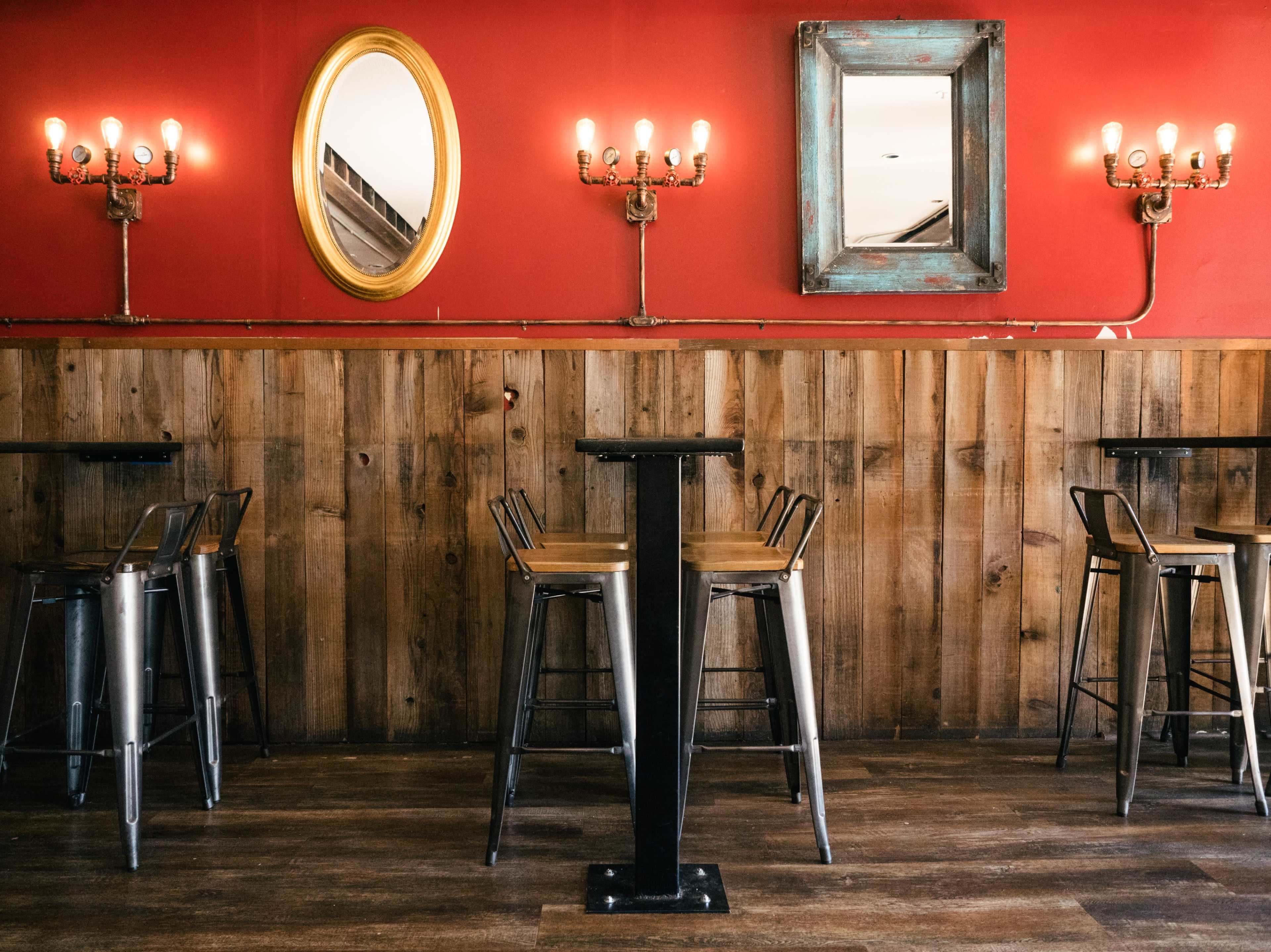 The image shows a rustic interior of a restaurant featuring wooden walls, metal bar stools, and ornate mirrors on a red backdrop.