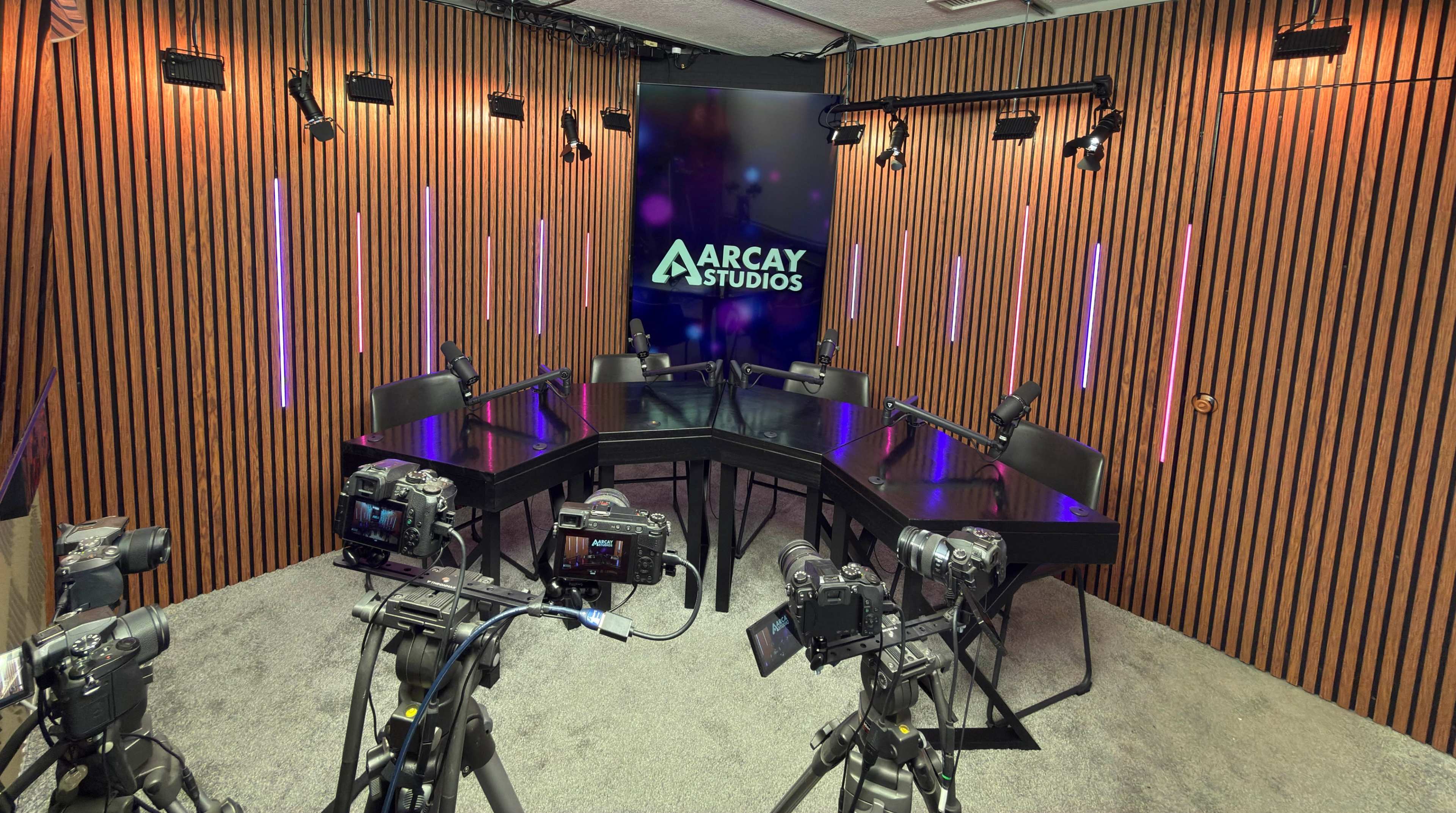 The image depicts a modern studio setup with a large, black table surrounded by chairs, multiple cameras on tripods facing the table, and wooden paneling in the background with colorful lighting.