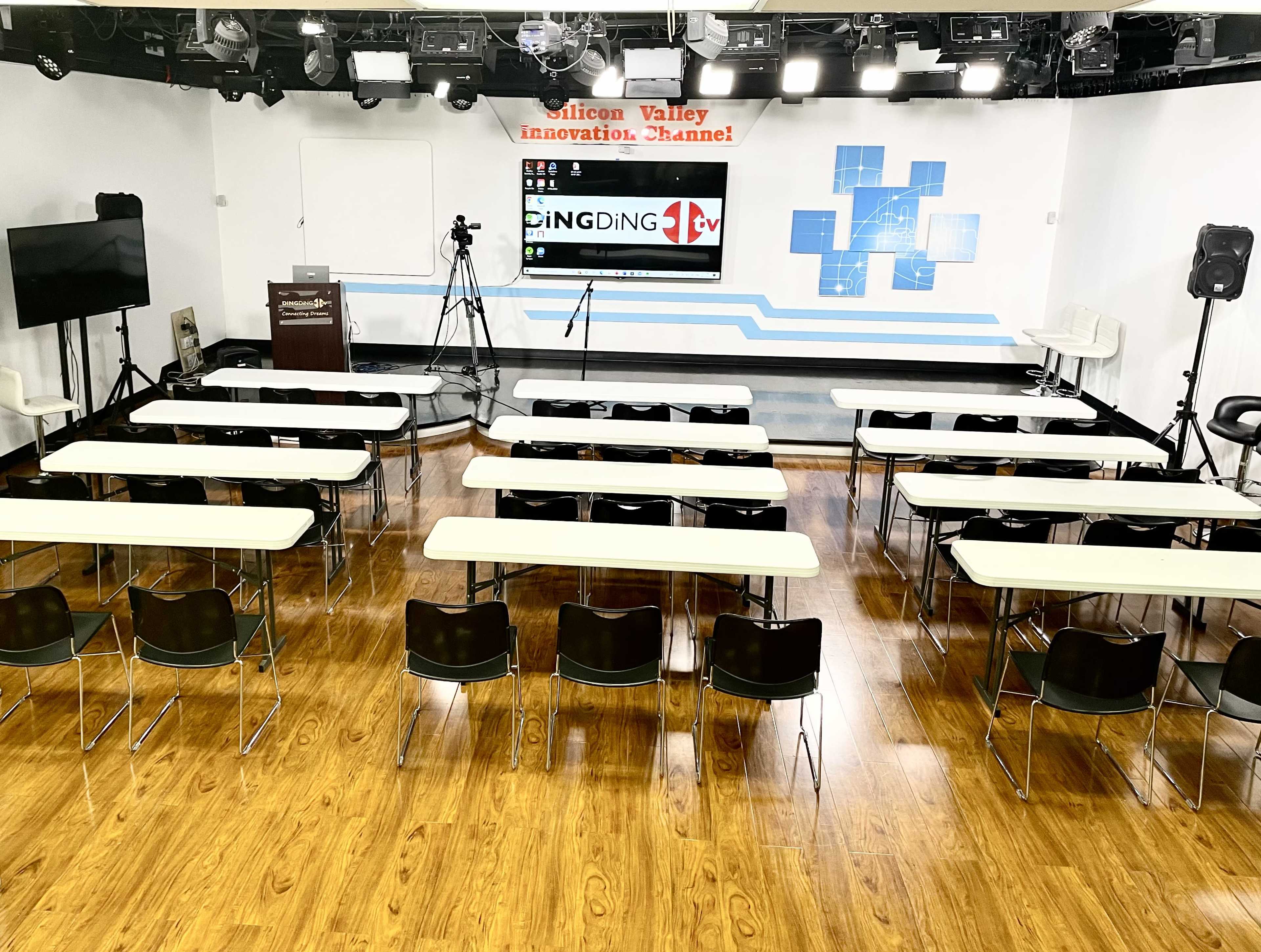 A conference room setup with rows of tables and chairs facing a screen, along with camera equipment and studio lights.